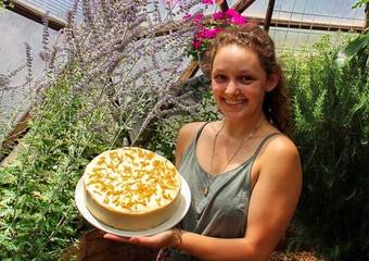 Gardener holding a frosted carrot cake among flowering plants inside a greenhouse