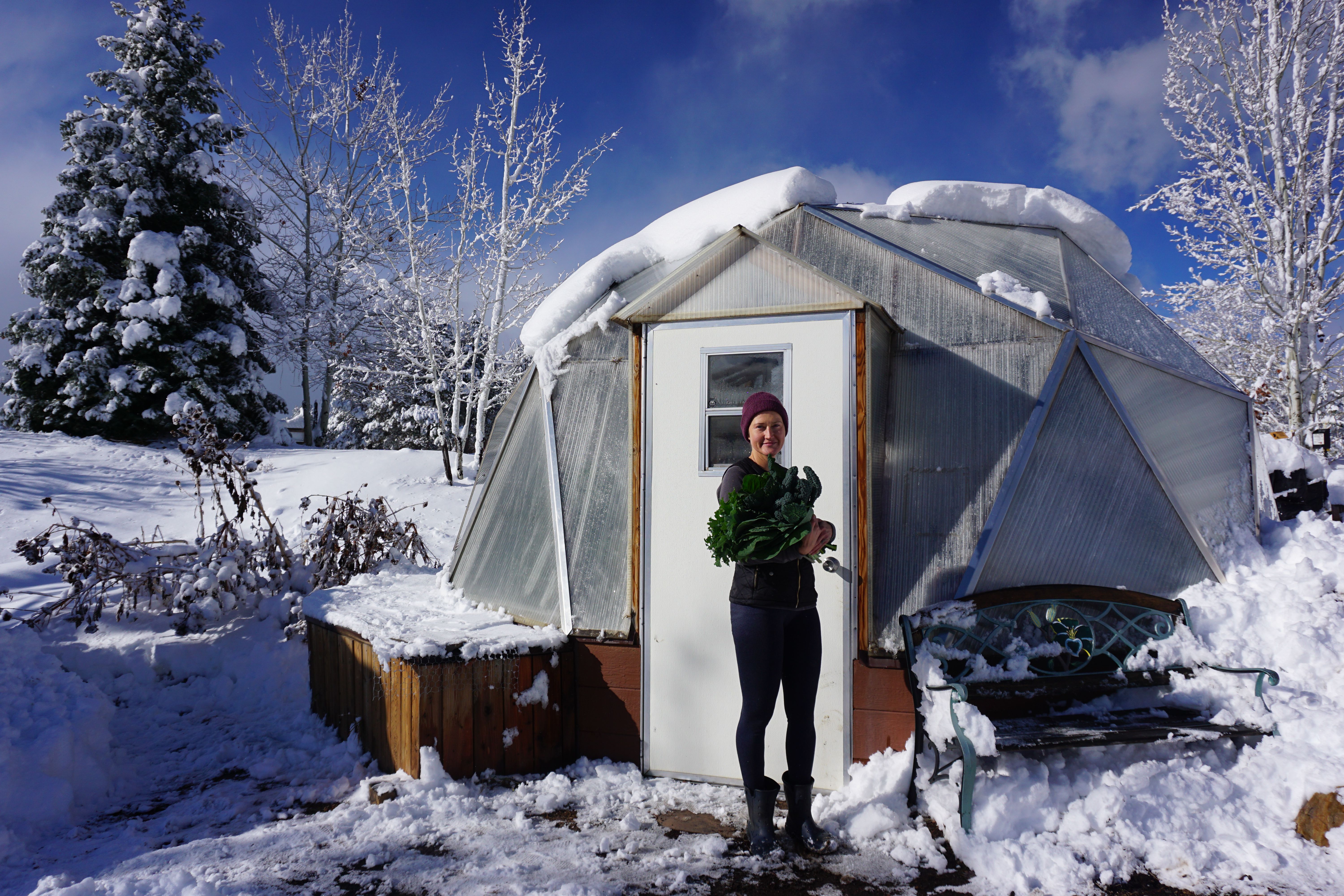 Gardener standing outside of her snow covered year-round greenhouse holding a freshly picked winter vegetable harvest
