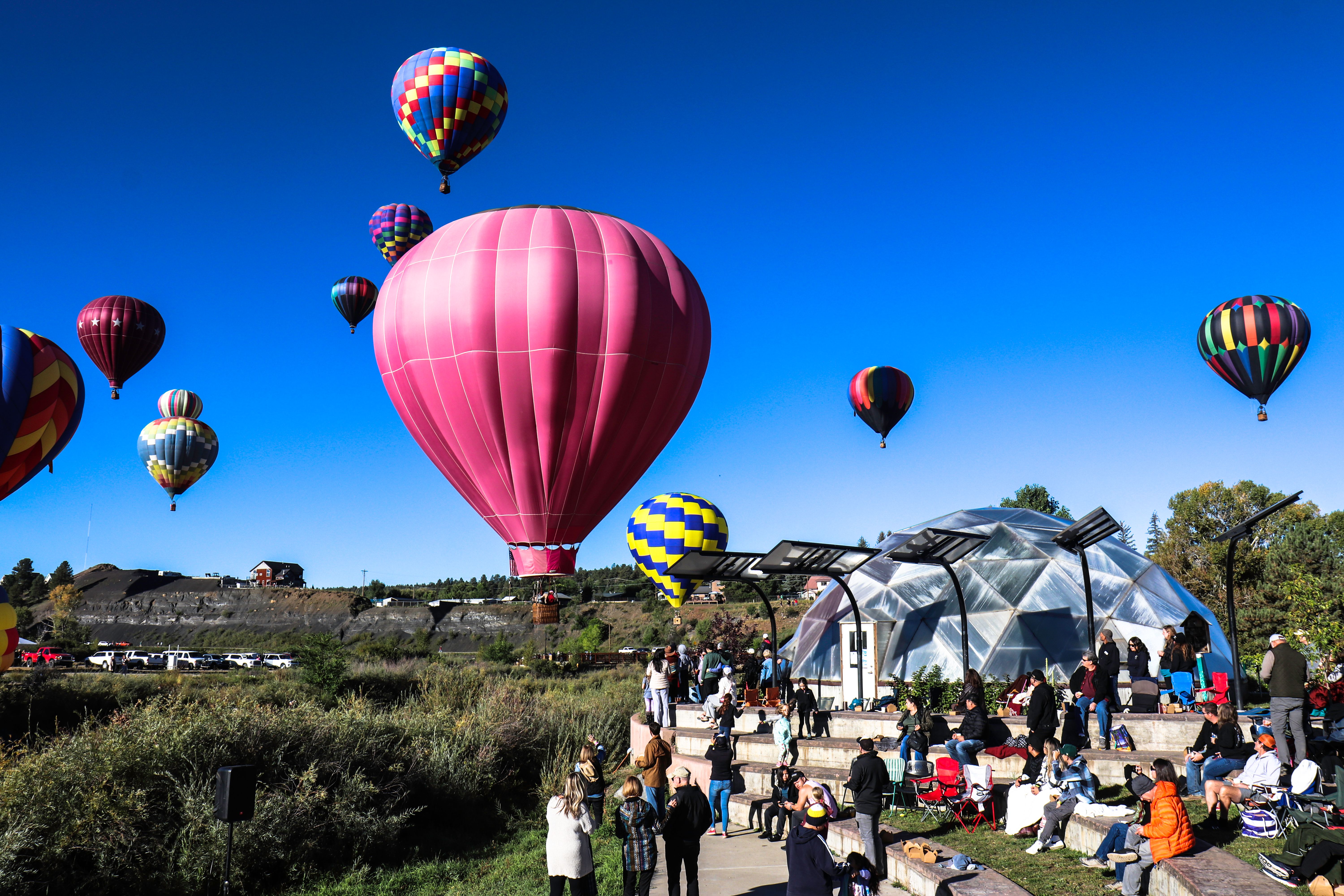 Hot air balloons in flights surrounding a Growing Dome greenhouse with a crowd of community members outside