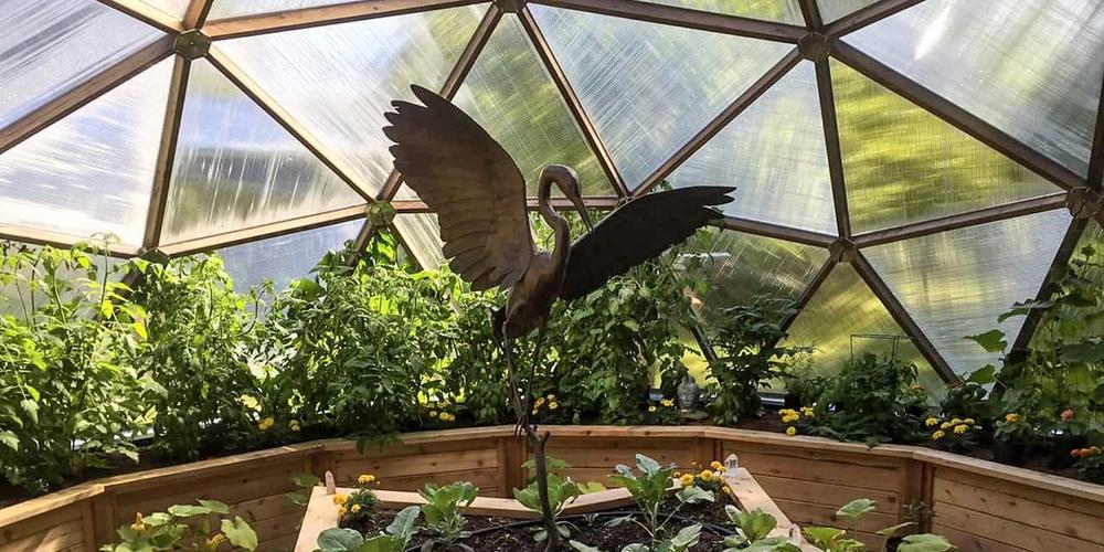 sun shining through polycarbonate panels of a growing dome greenhouse with a large crane sculpture in the middle bed