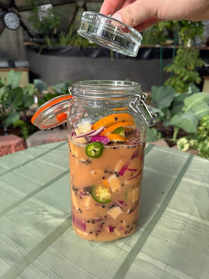 A large glass jar filled with fire cider ingredients on a table with a light green tablecloth being topped with a glass weight