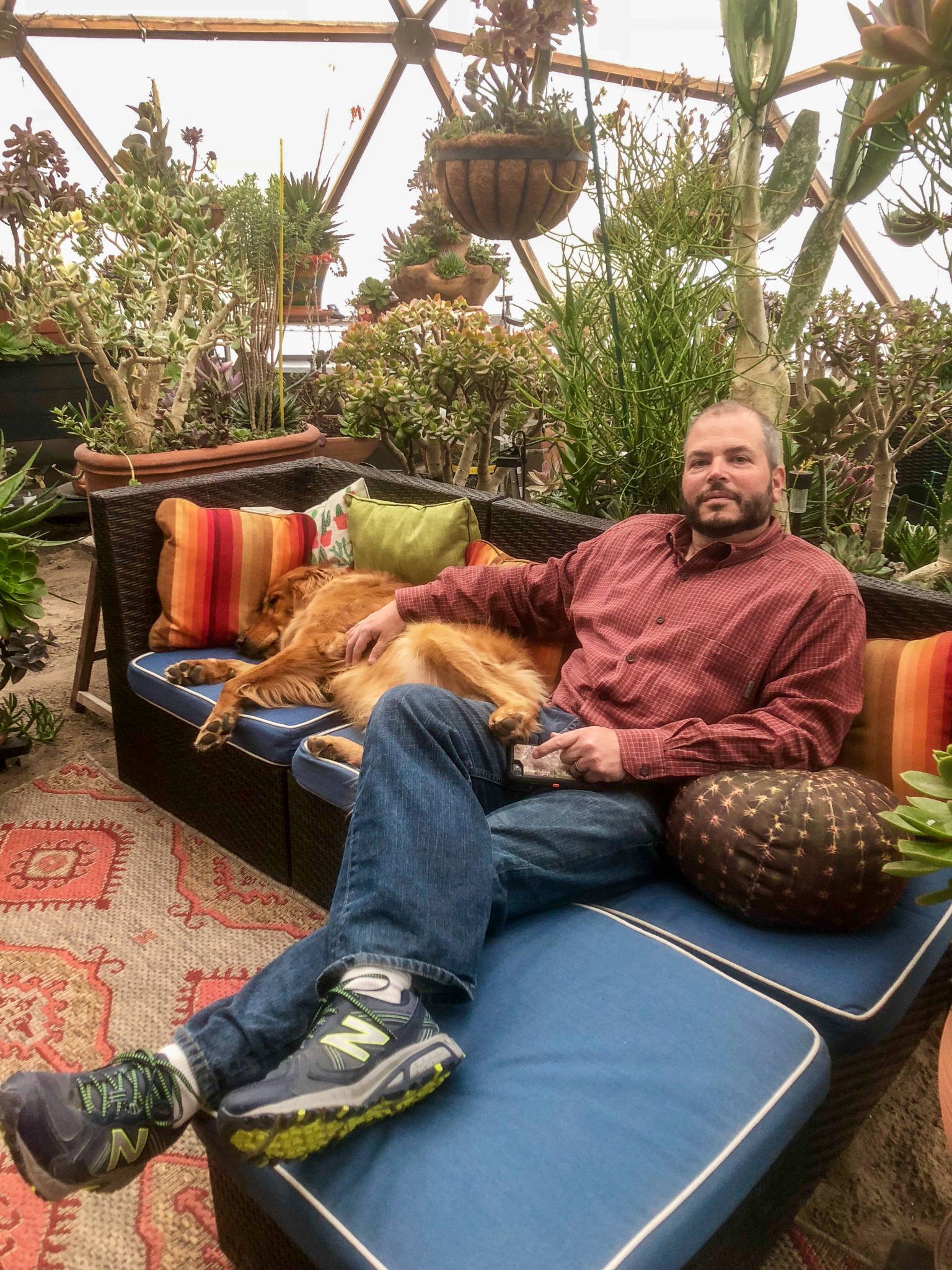 man sitting with a dog relaxing on a couch inside a geodesic greenhouse with lots of plants