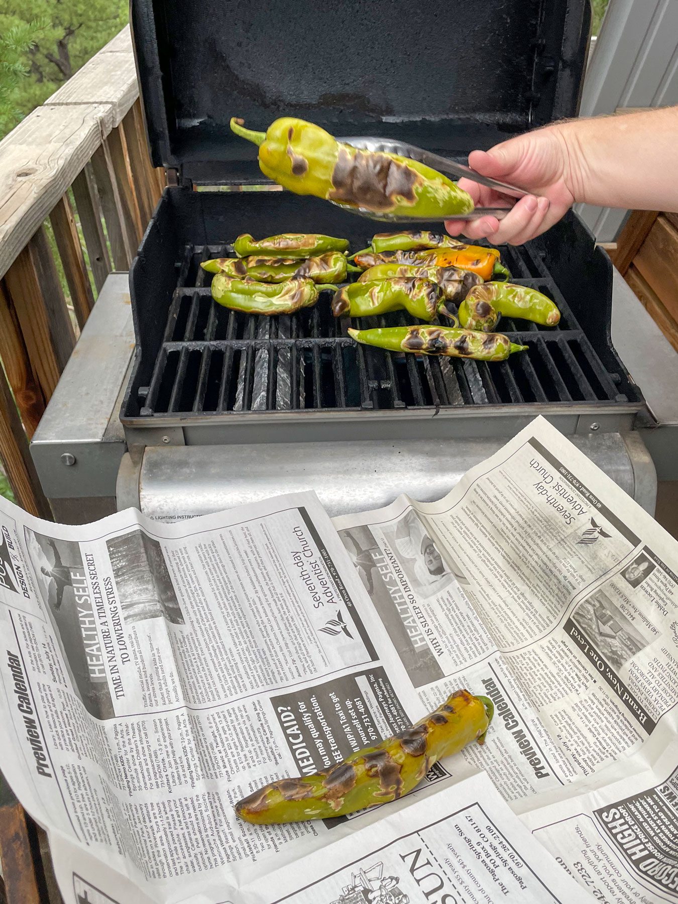 using tongs to remove roasted green chile from a grill and placing them on newspaper