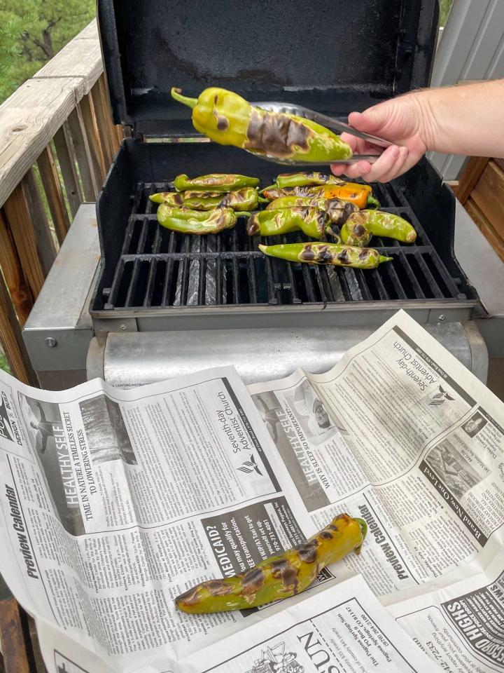 using tongs to remove roasted green chile from a grill and placing them on newspaper