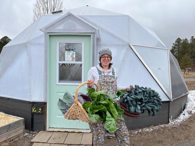 Gardener in winter hat holding two large baskets of freshly harvested vegetables from her year-round greenhouse