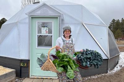 Woman holding two baskets full of greens outside her greenhouse in the winter