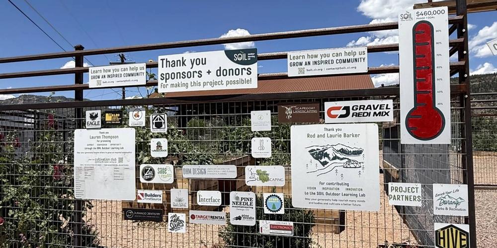 Signs from various businesses and donors on a metal fence surrounding a community garden