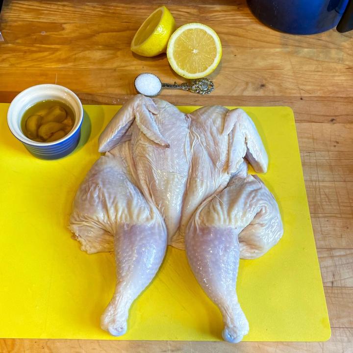 garlic in oil in a blue ramekin, a raw chicken on a plastic yellow cutting board, lemon halved, and a teaspoon of kosher salt on a butcher block