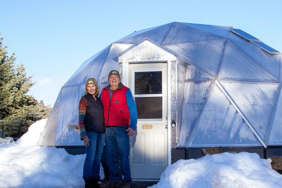 two people stand by the front door of a geodesic greenhouse with snow on the ground