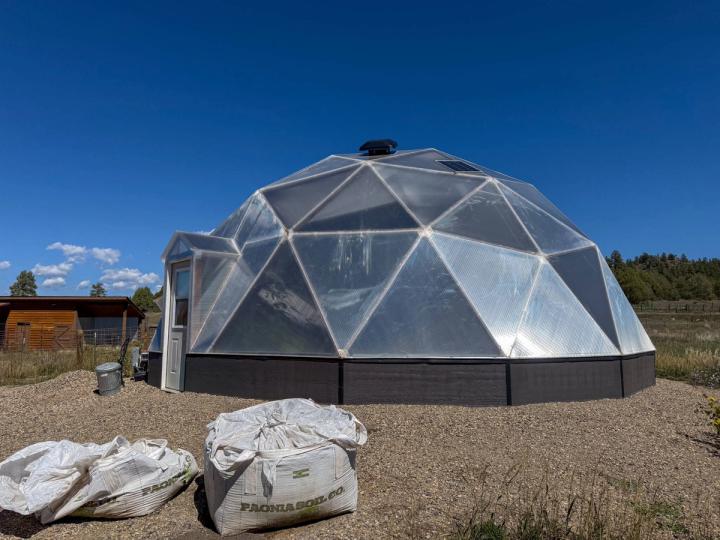 Exterior of a 33' geodesic growing dome greenhouse under blue sky with 2 bags of soil in foreground