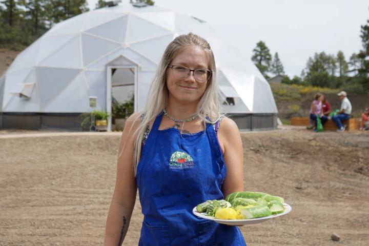 lady holding vegetables on a tray in front of a greenhouse