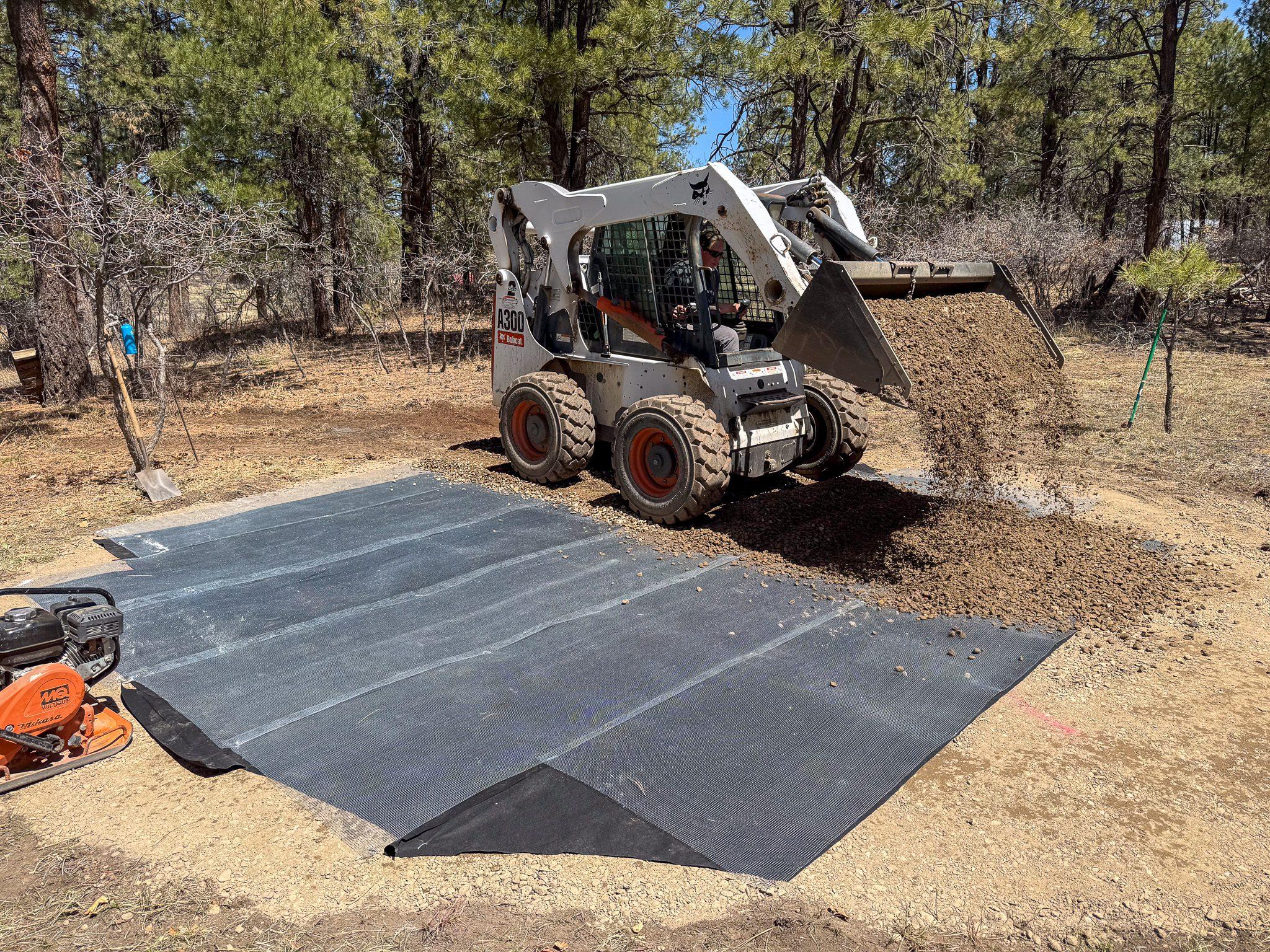 A skid steer placing gravel over weed barrier during greenhouse site preparation