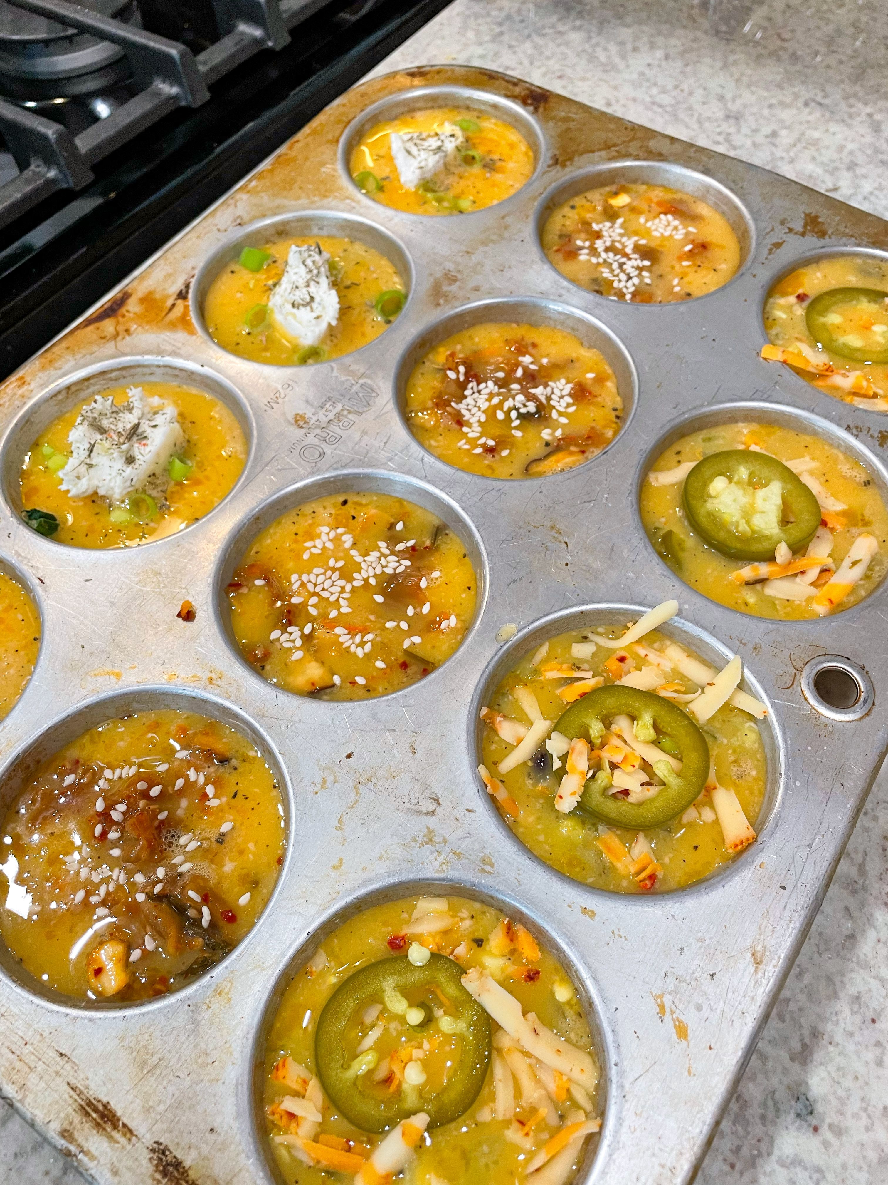 egg bites ready for the oven topped with goat cheese sesame seeds or jalapenos and cheese