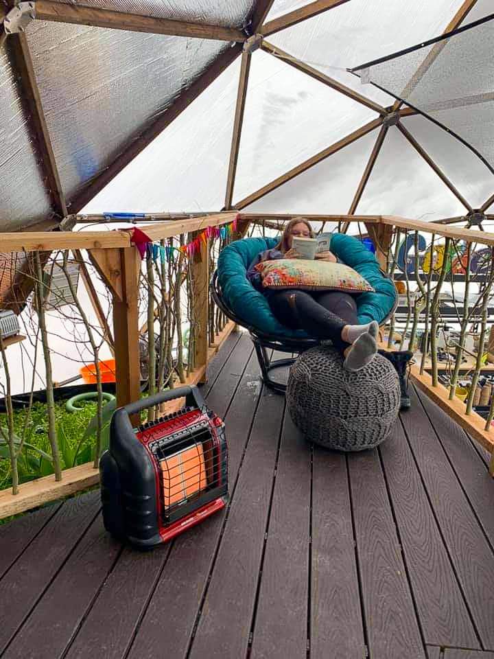 person sitting in a chair on a deck with a propane heater above the pond inside the geodesic greenhouse