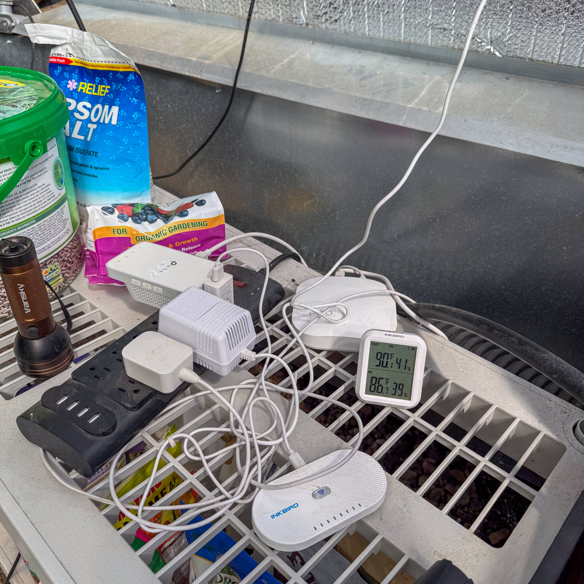 Various sensors and Wifi adaptors resting on a plastic shelf with a thermometer and garden amendments around them