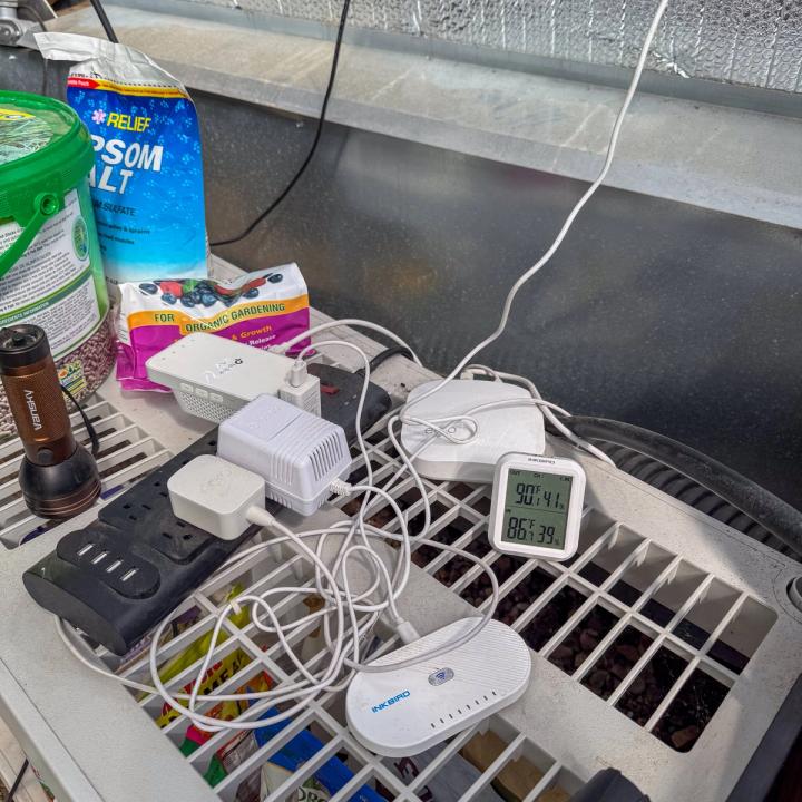 Various sensors and Wifi adaptors resting on a plastic shelf with a thermometer and garden amendments around them