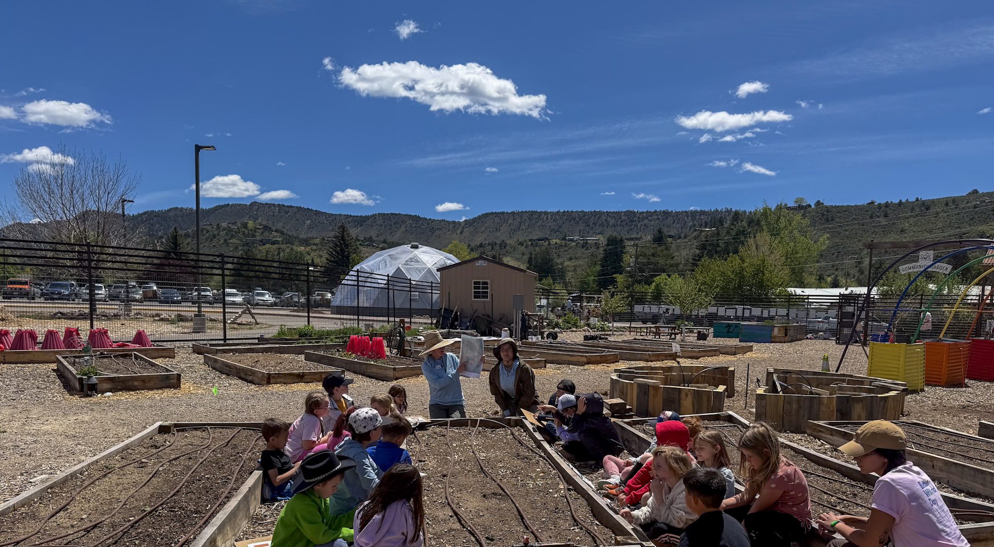 Students seated around an outdoor raised garden bed with a teacher instructing them. Growing Dome seen in the background.
