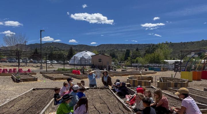 Students seated around an outdoor raised garden bed with a teacher instructing them. Growing Dome seen in the background.