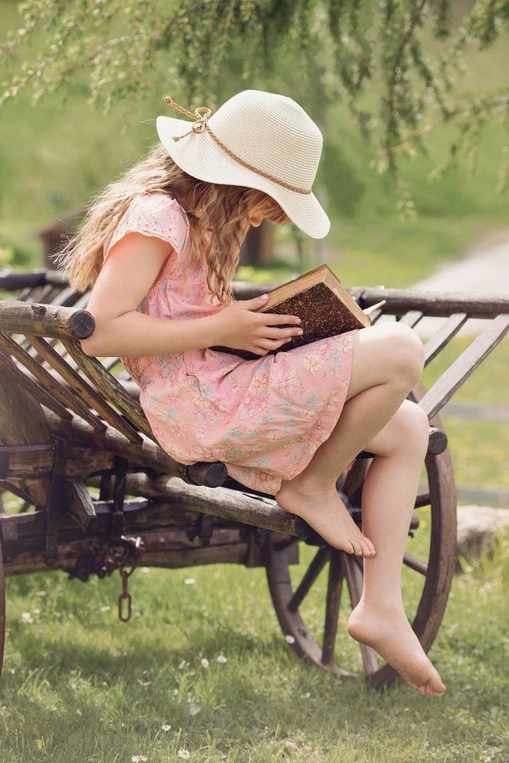 girl sitting outside on a cart barefoot with a pink dress and sun hat on and reading a book