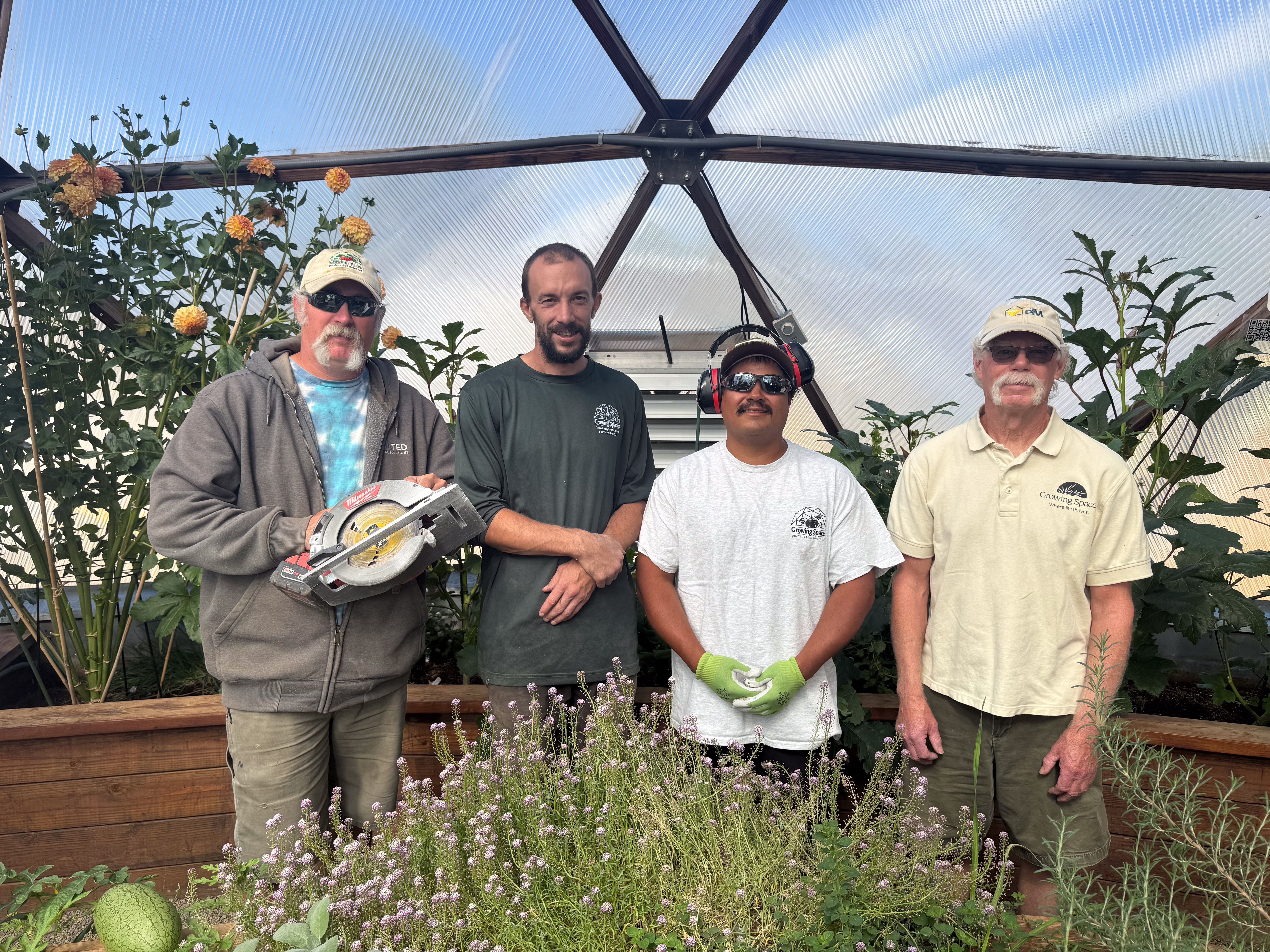 Four people standing together inside a Growing Dome greenhouse, one holding a saw