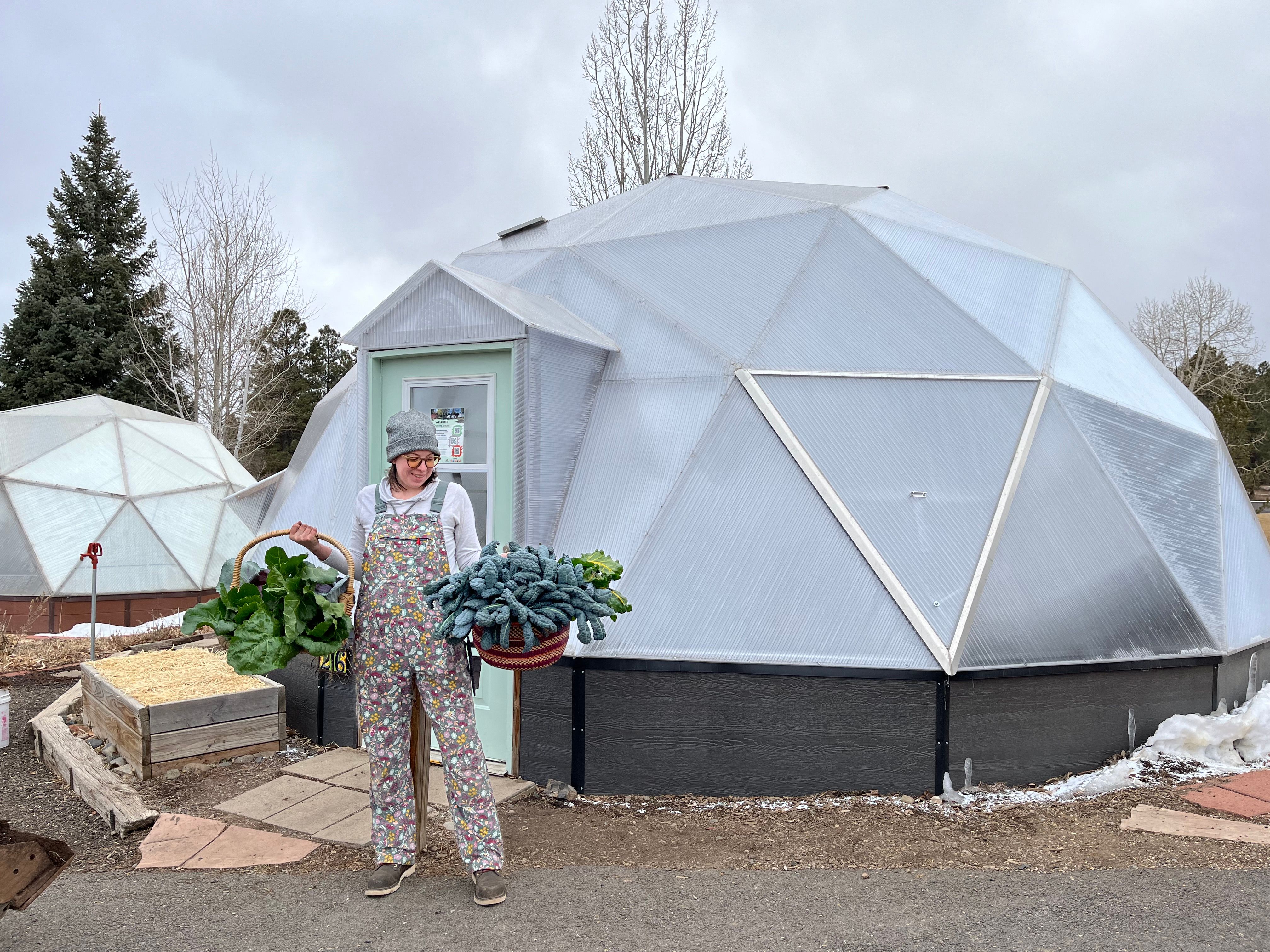 Woman holding two baskets full of leafy greens standing outside the door of a Growing Dome greenhouse