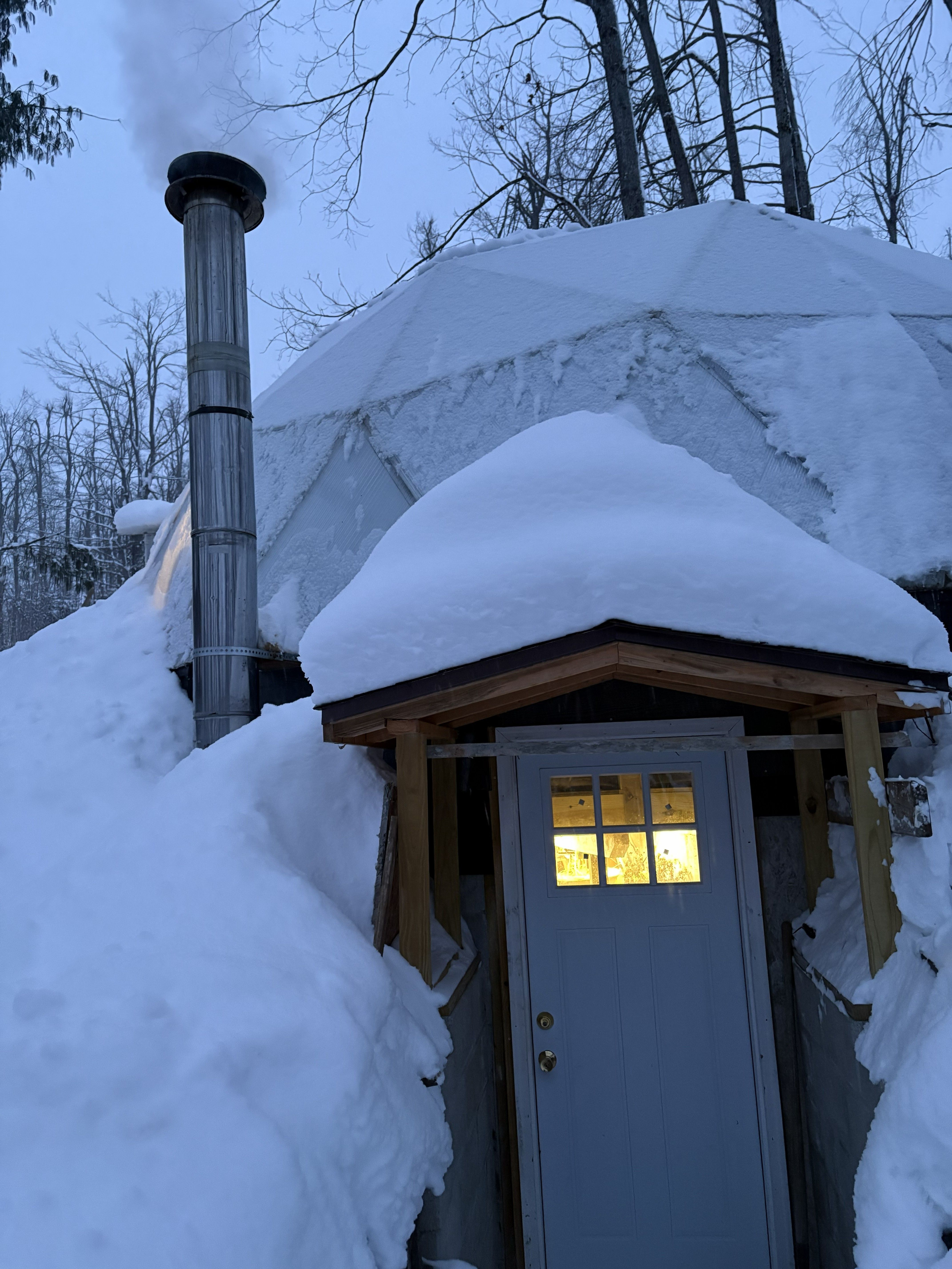 A snow-covered greenhouse with a custom basement entrance and wood-stove chimney