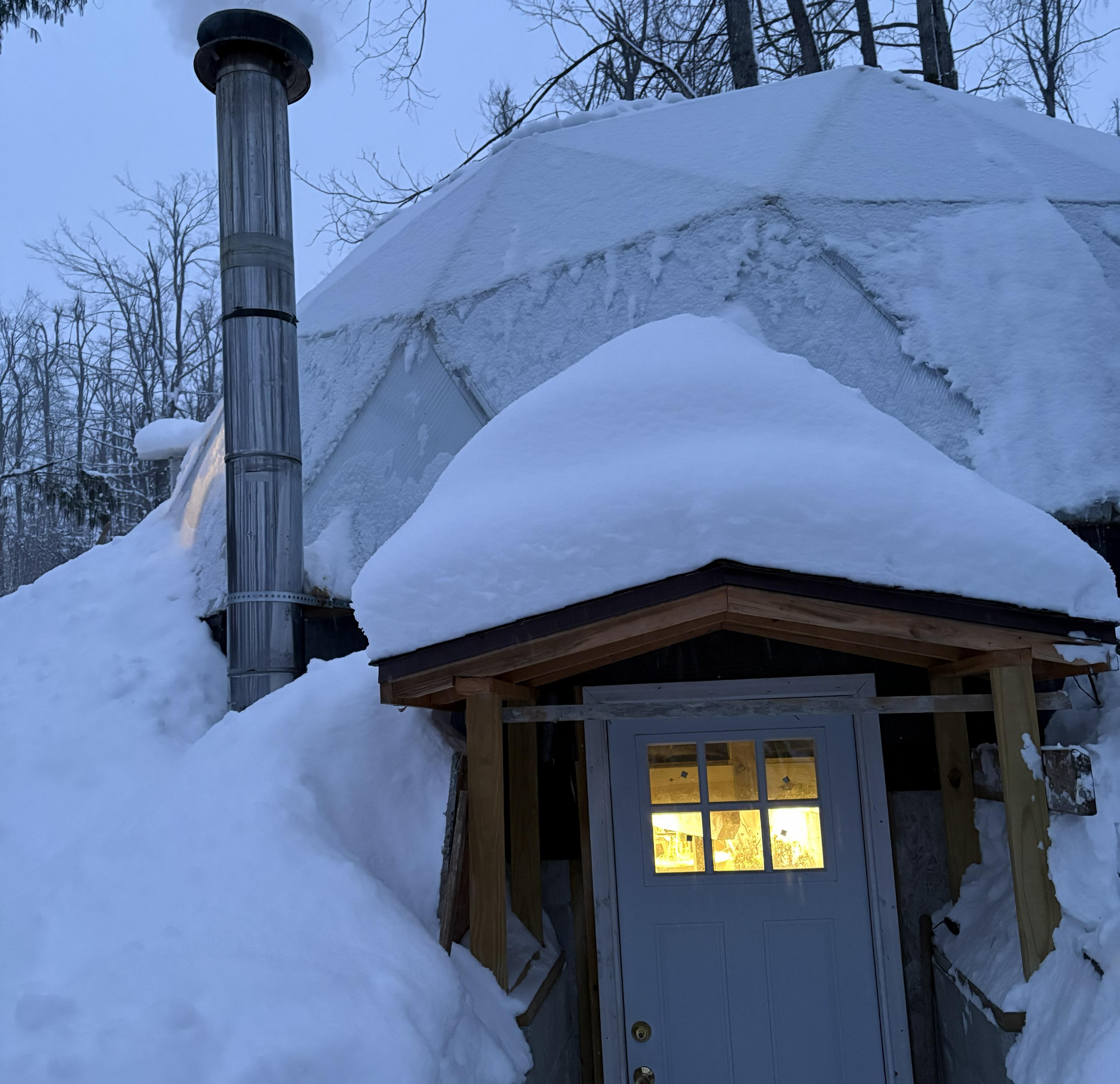 A snow-covered greenhouse with a custom basement entrance and wood-stove chimney