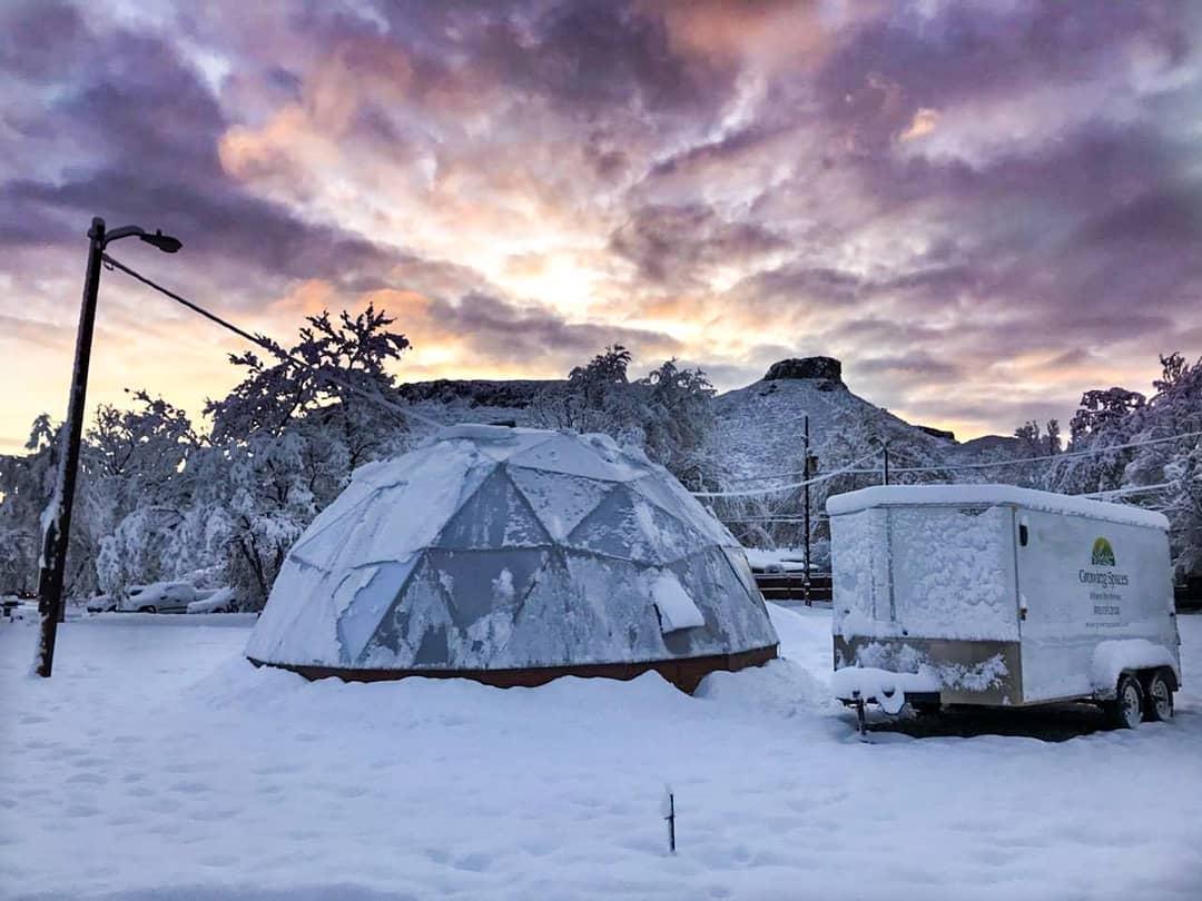 Growing dome greenhouse and growing spaces trailer covered in a light layer of snow under a dramatic purple sunset