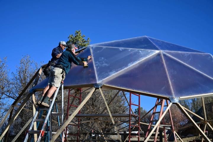 Two men on ladders attaching polycarbonate panels to a geodesic greenhouse with screws
