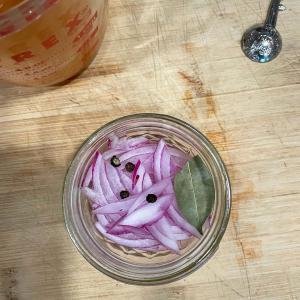 thinly sliced red onion with peppercorns and bay leaf in jar with measuring cup of brine on cutting board