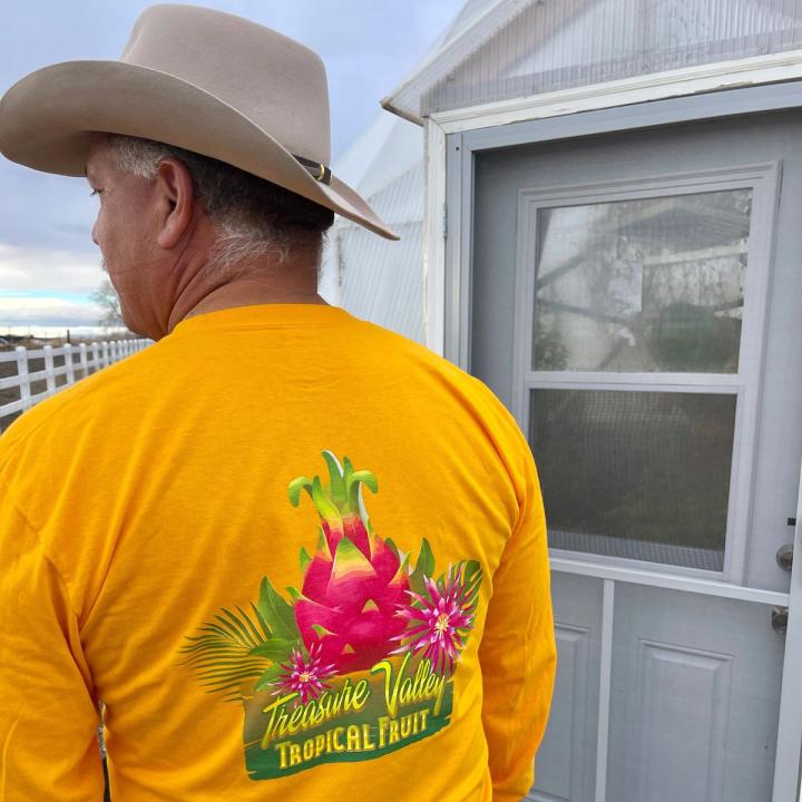 A man wearing a bright yellow shirt with a logo on the back of a tropical dragon fruit that says "Treasure Valley Tropical Fruit." He's standing at the door to his Growing Dome.