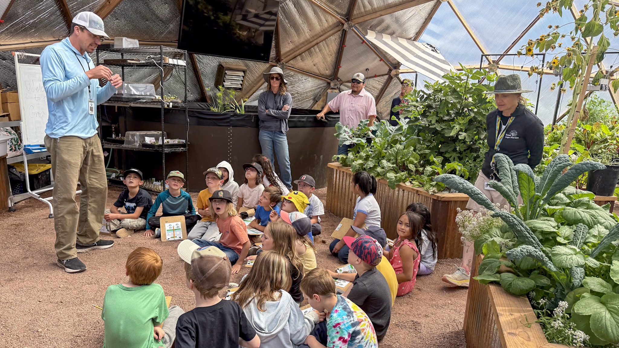students parents and facilitators learning inside of a growing dome greenhouse with lots of vegetables growing in the raised beds