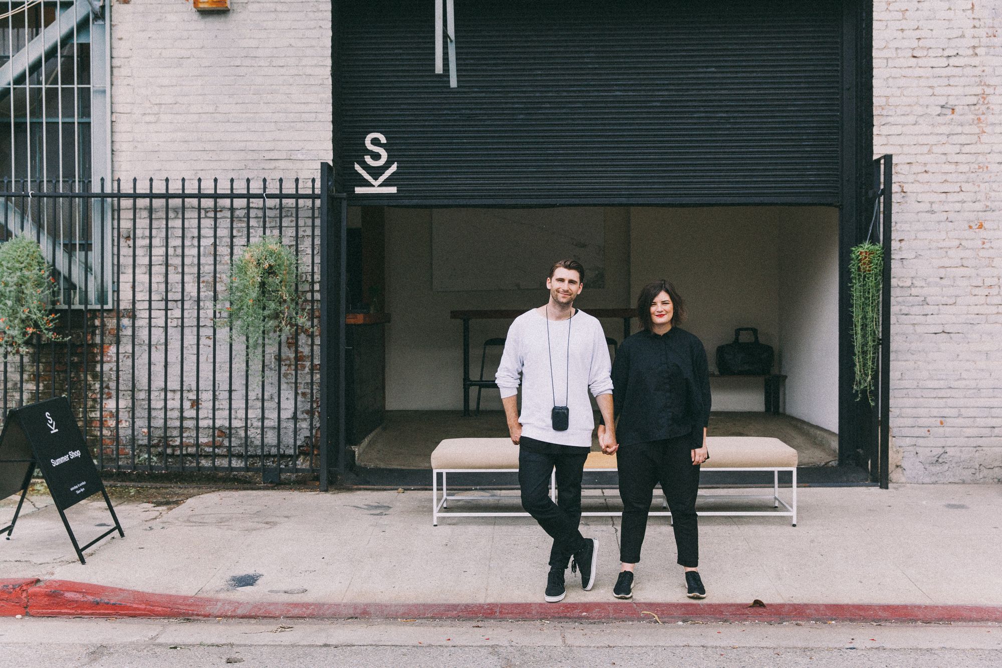 A man and woman holding hands stand in front of a modern storefront with a large black open garage door and an SK logo, with a Summer Shop sign nearby.