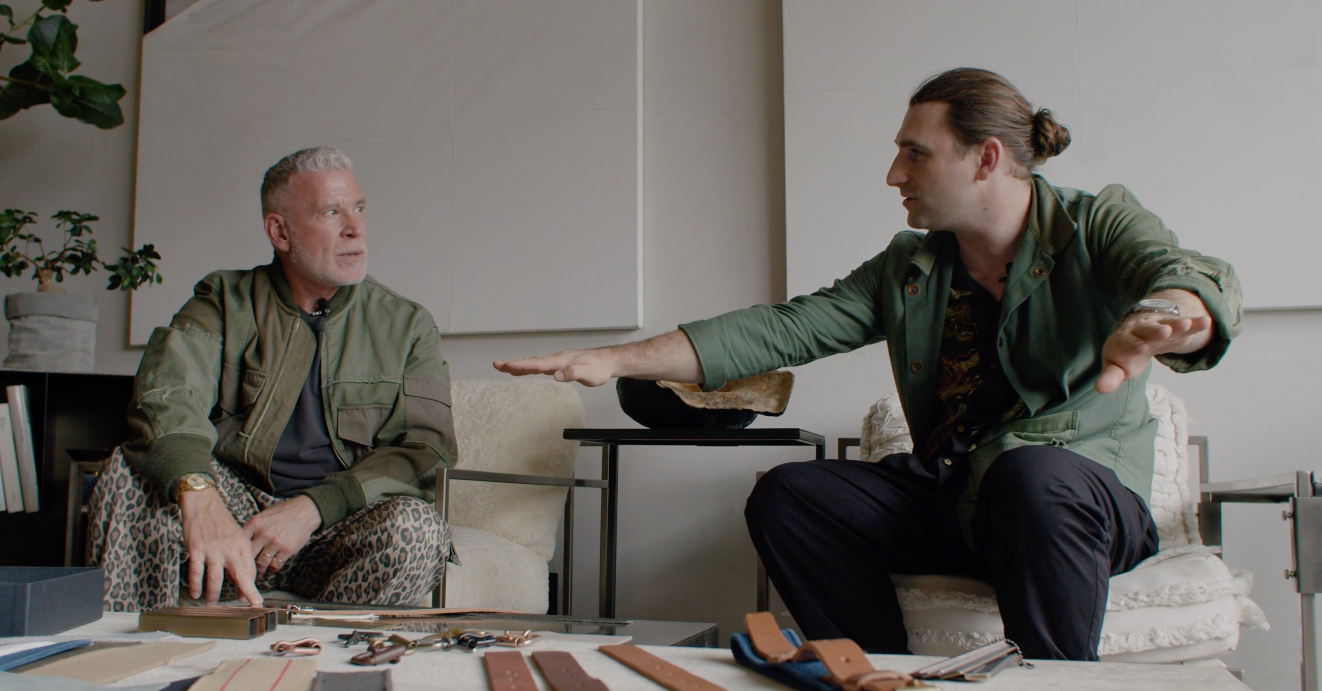 Two men sit at a table covered with leather watch straps; one with grey hair, the other with a bun, both wearing green jackets and talking.