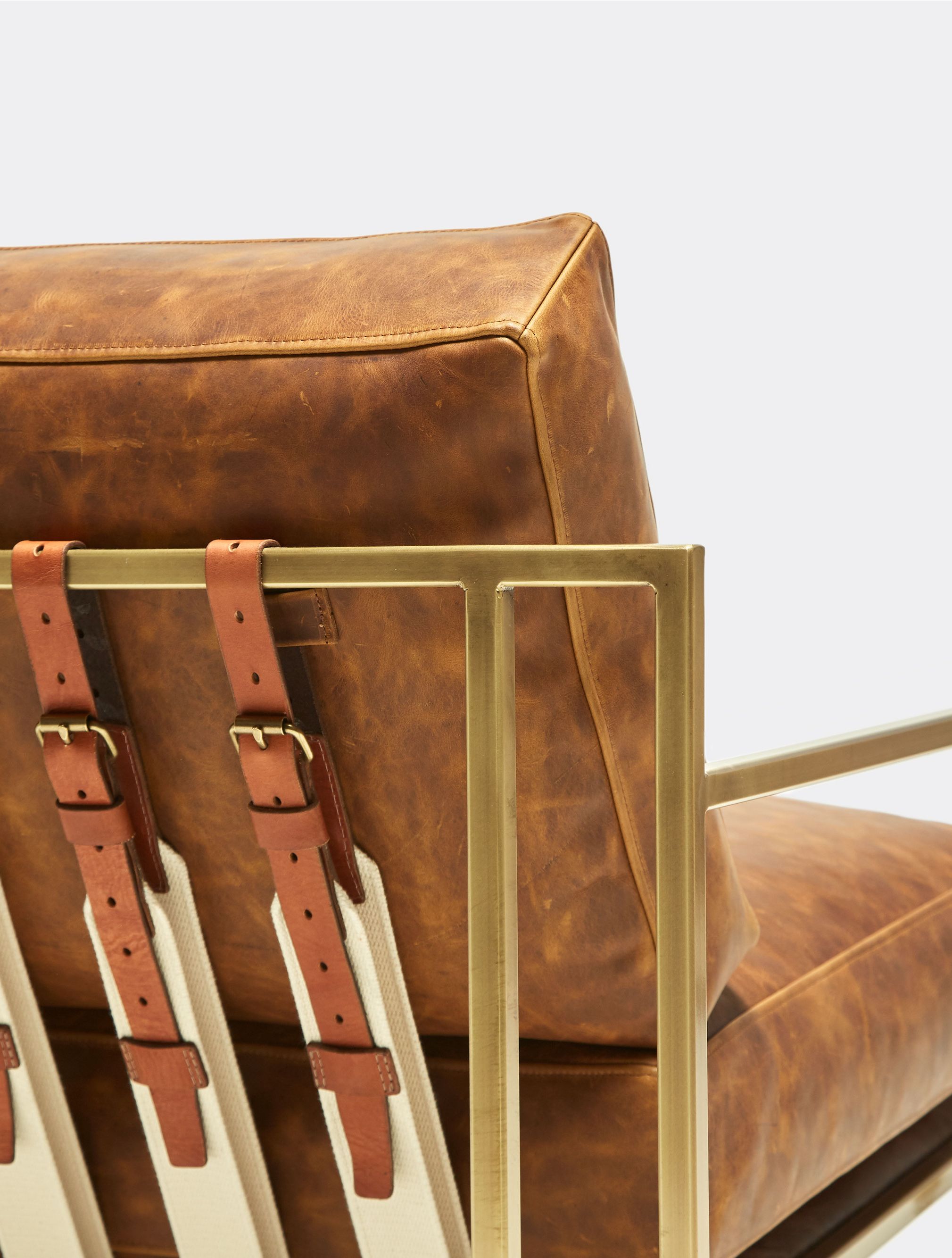 a brown leather chair with leather and webbing straps and a brass finished steel frame on a white background .