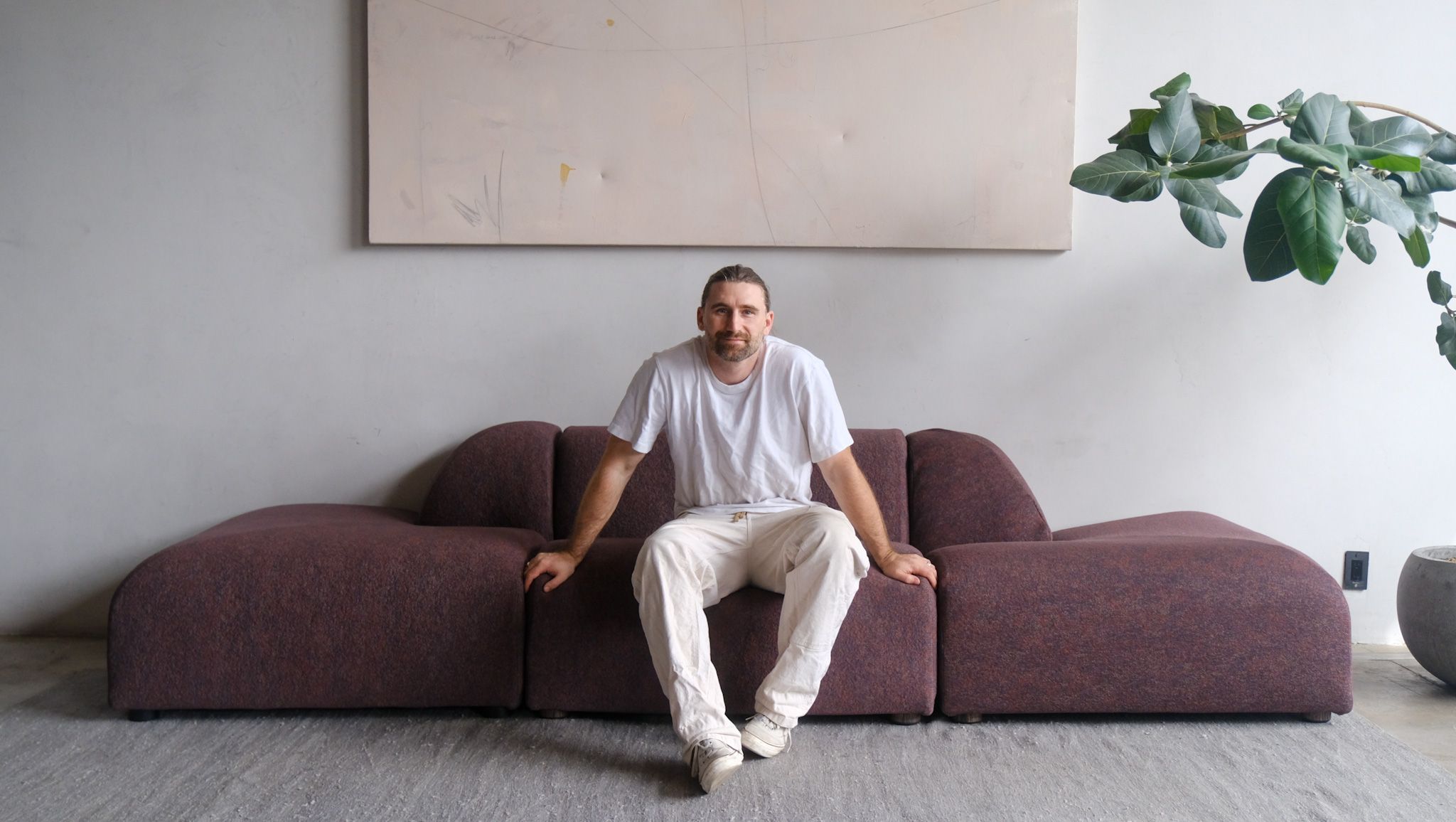 Man with beard and ponytail sitting on a dark red modular sofa against a grey wall with an abstract painting.