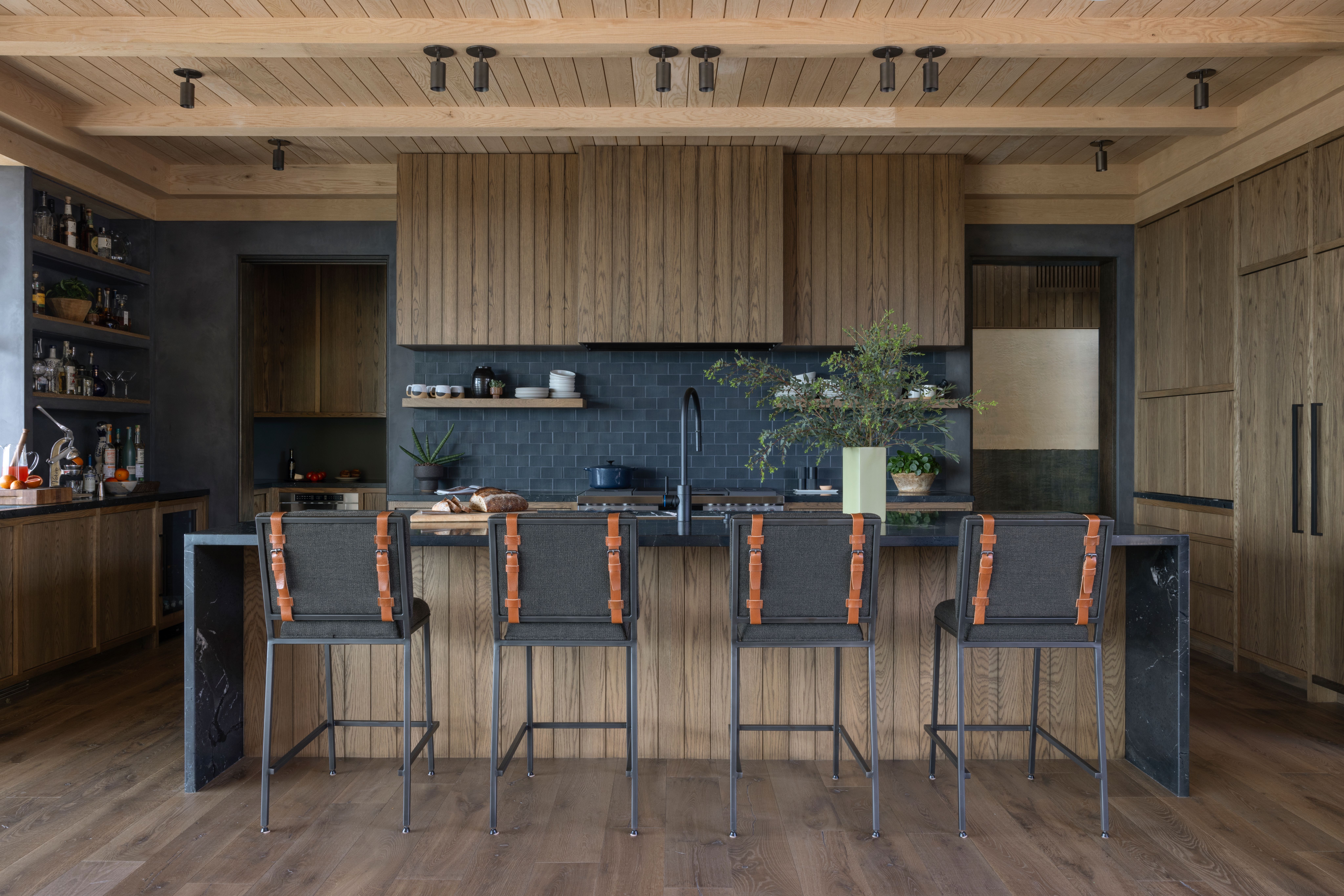 A modern kitchen with natural wood cabinetry, a dark subway tile backsplash, and a wooden island with four bar stools.