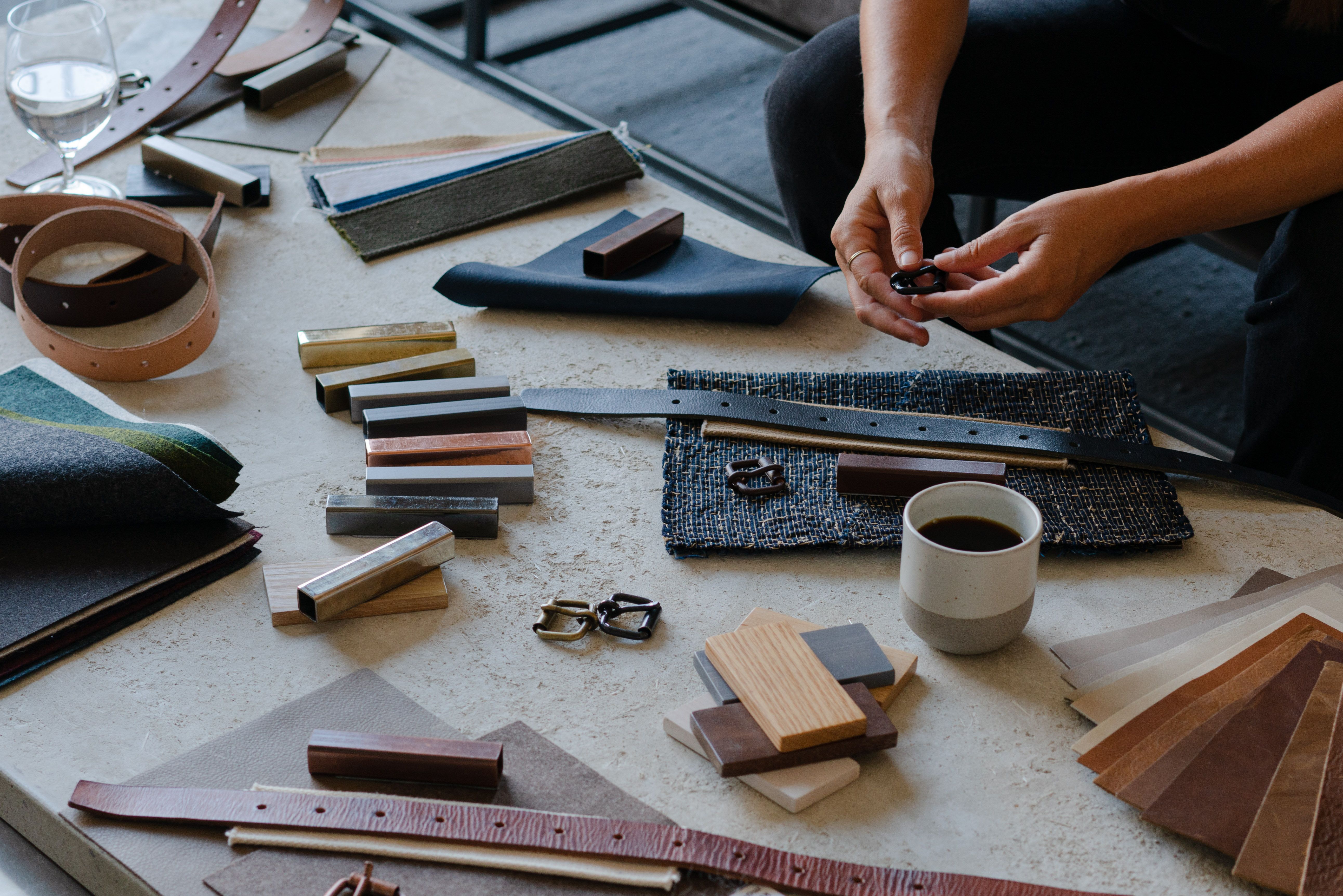 Hands examine a buckle amidst a variety of design materials like leather, fabric, wood, and metal on a table.