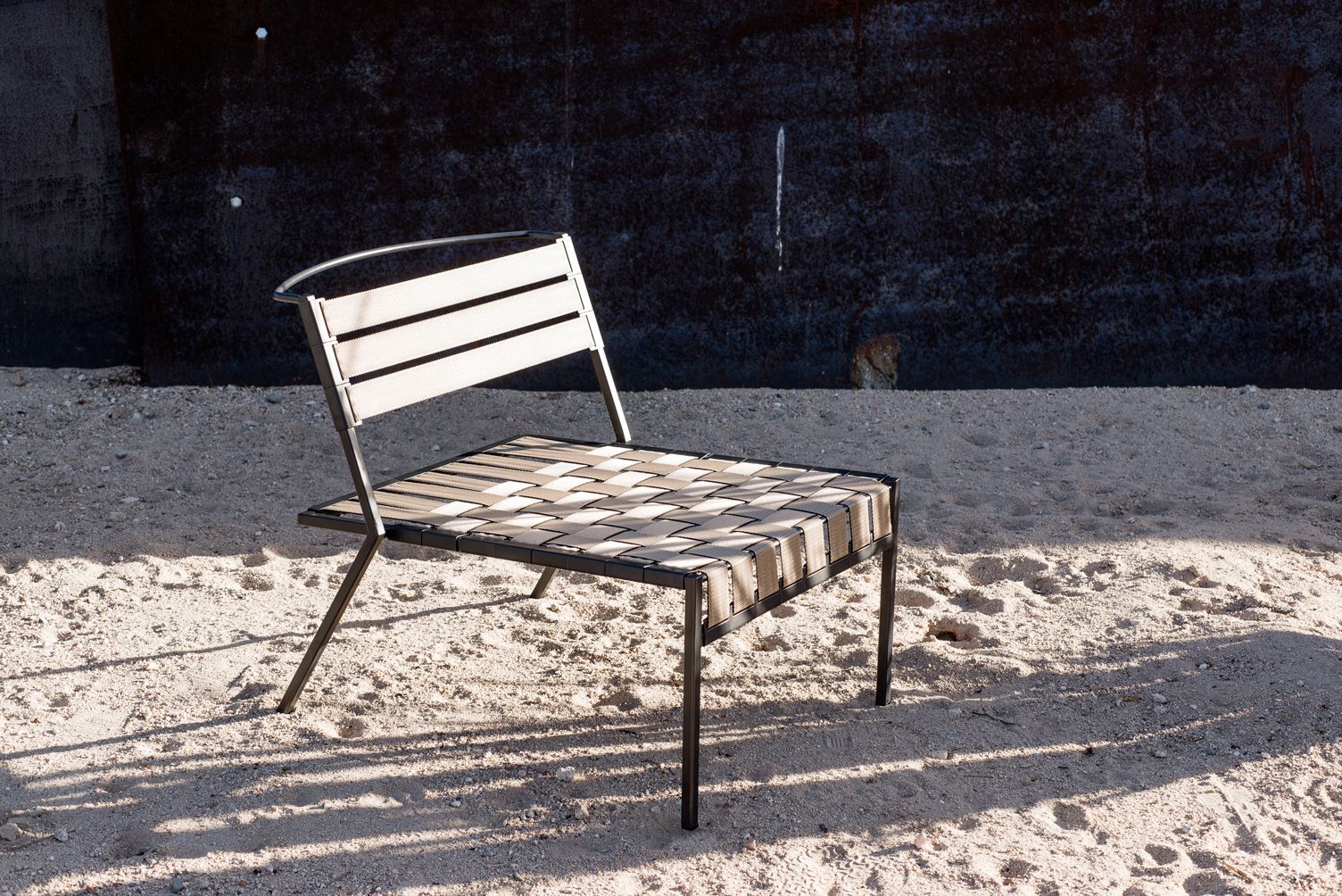 An outdoor chair with a slatted back and woven seat sits on sand in front of a dark wall.