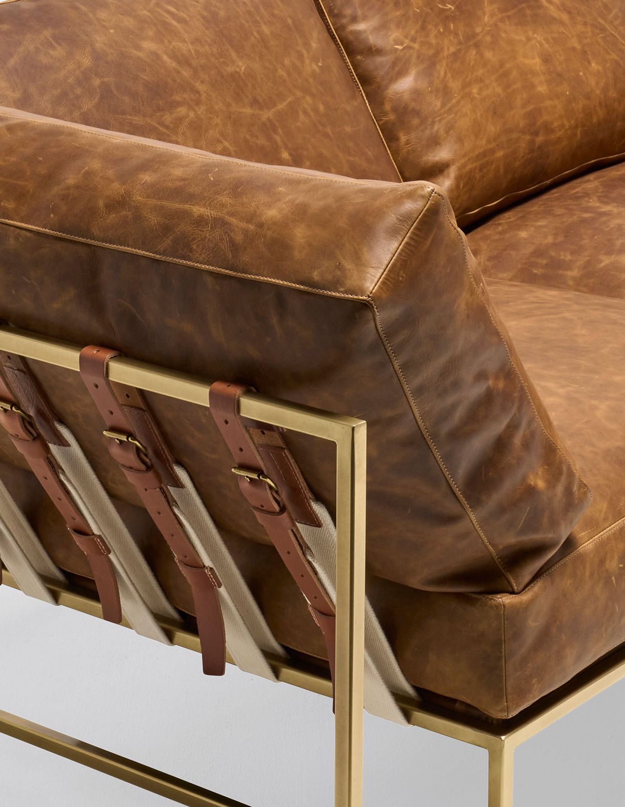 a brown leather sectional sofa with a brass finished steel frame and leather and webbing supporting belts shown on a white background .