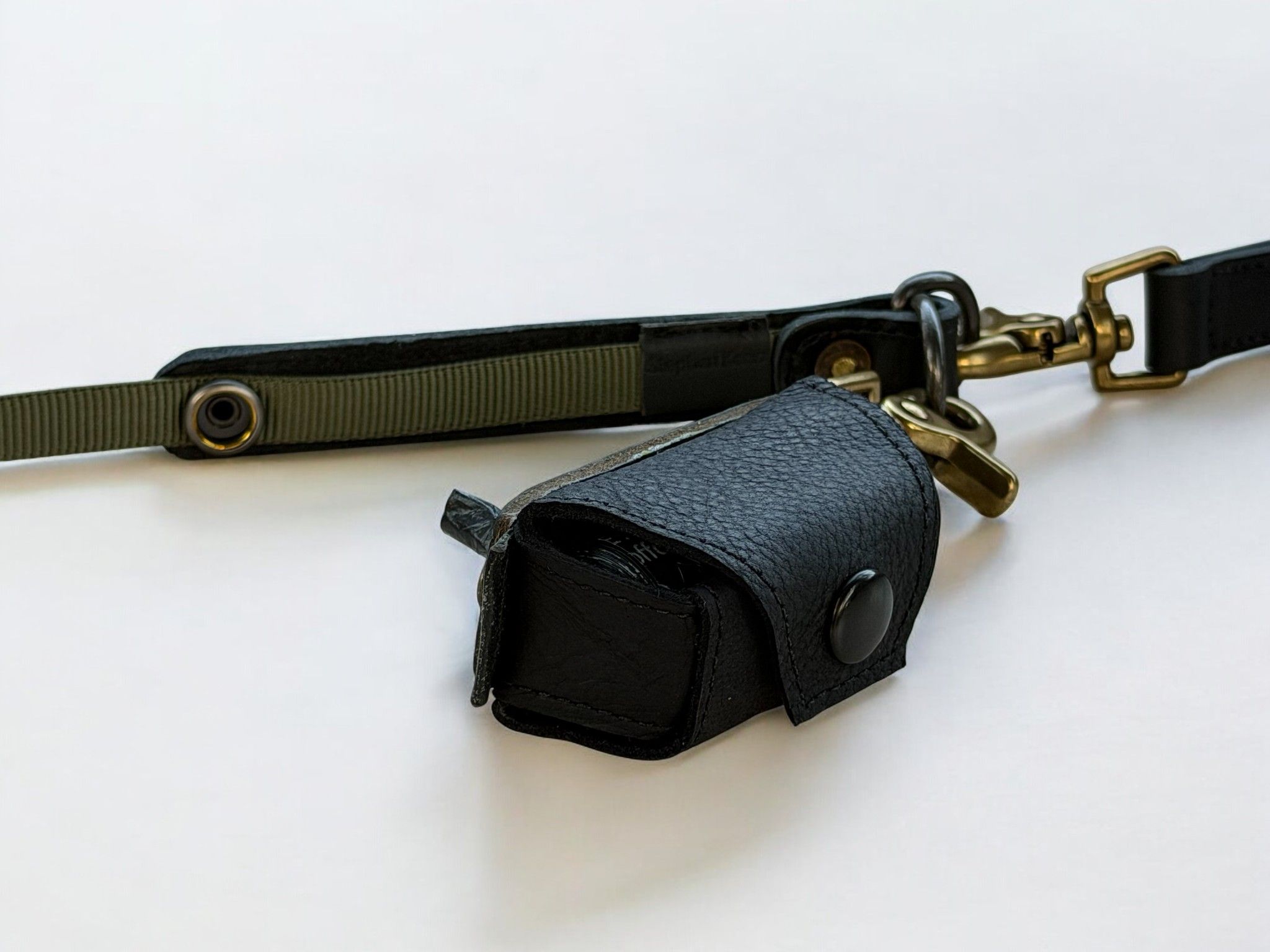 A black leather poop bag holder with olive green webbing and brass hardware, with an attached small black leather snap-pouch.
