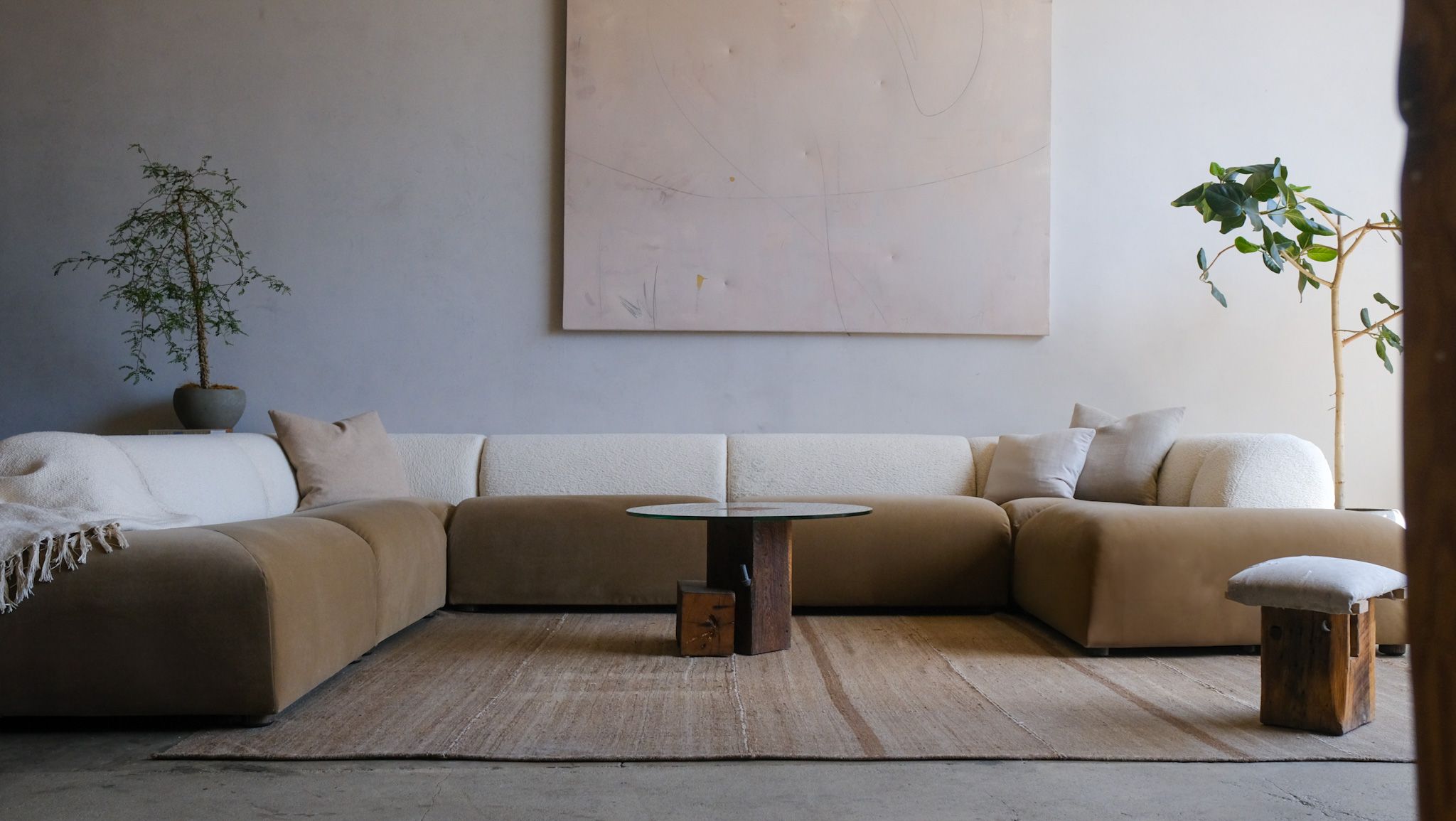 A modern living room featuring a two-toned tan and white modular sofa, a round glass coffee table, abstract wall art, and potted plants.