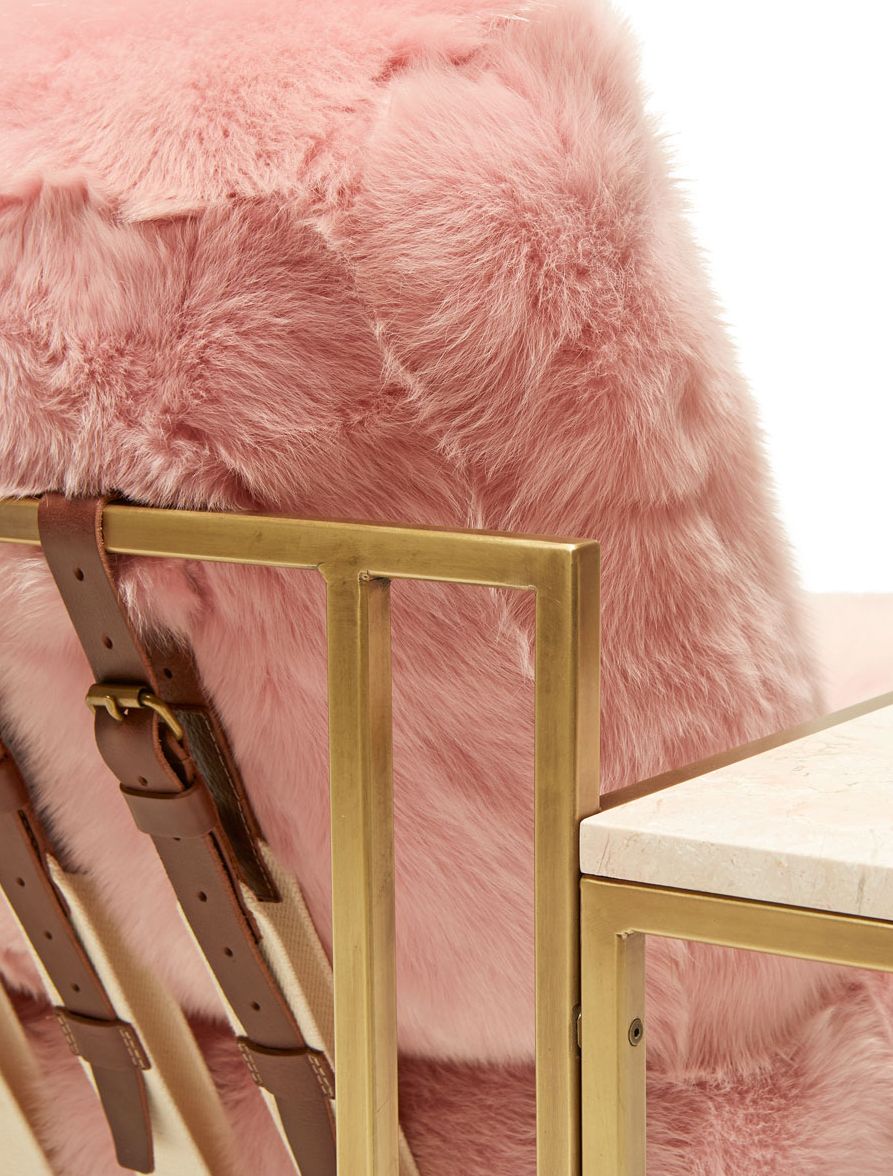 a pink shearling armchair with leather and webbing supporting belts on a brass finished steel frame and a pink marble side table attached shown on a white background .