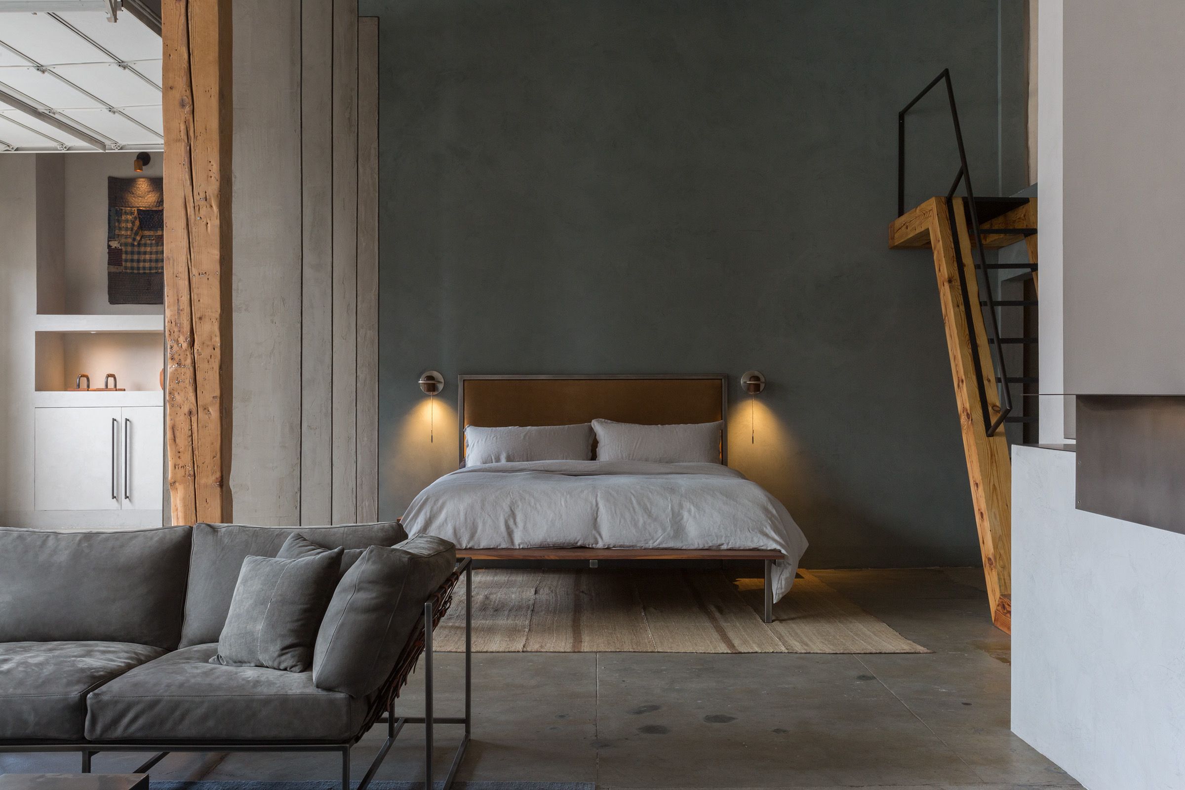 there is a large bed in the middle of a loft style apartment with wood pillars and a wood and steel staircase. A grey leather sofa floats in the center of the room