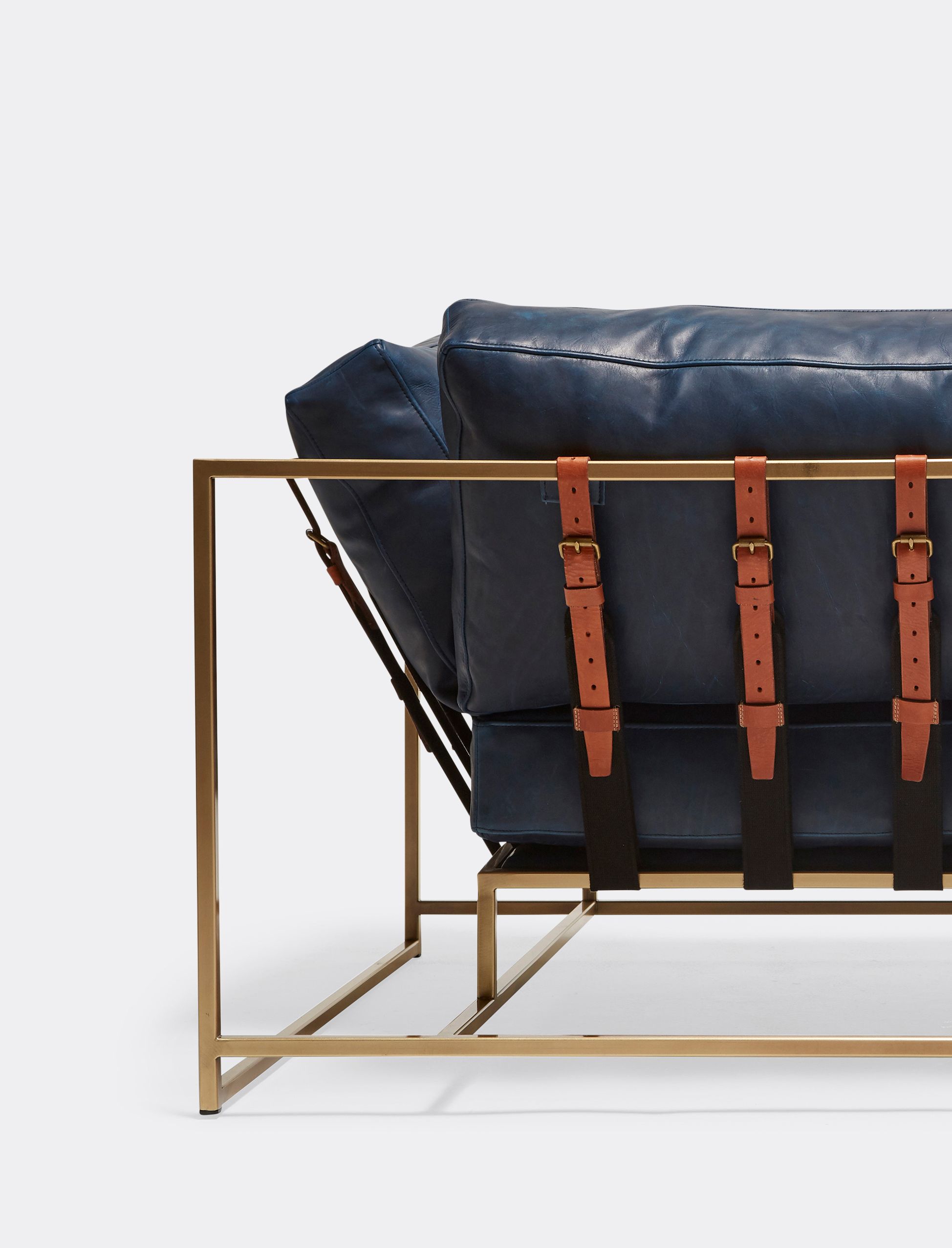 A dark blue leather couch with brown leather straps and a brass finished steel frame shown on a white background .