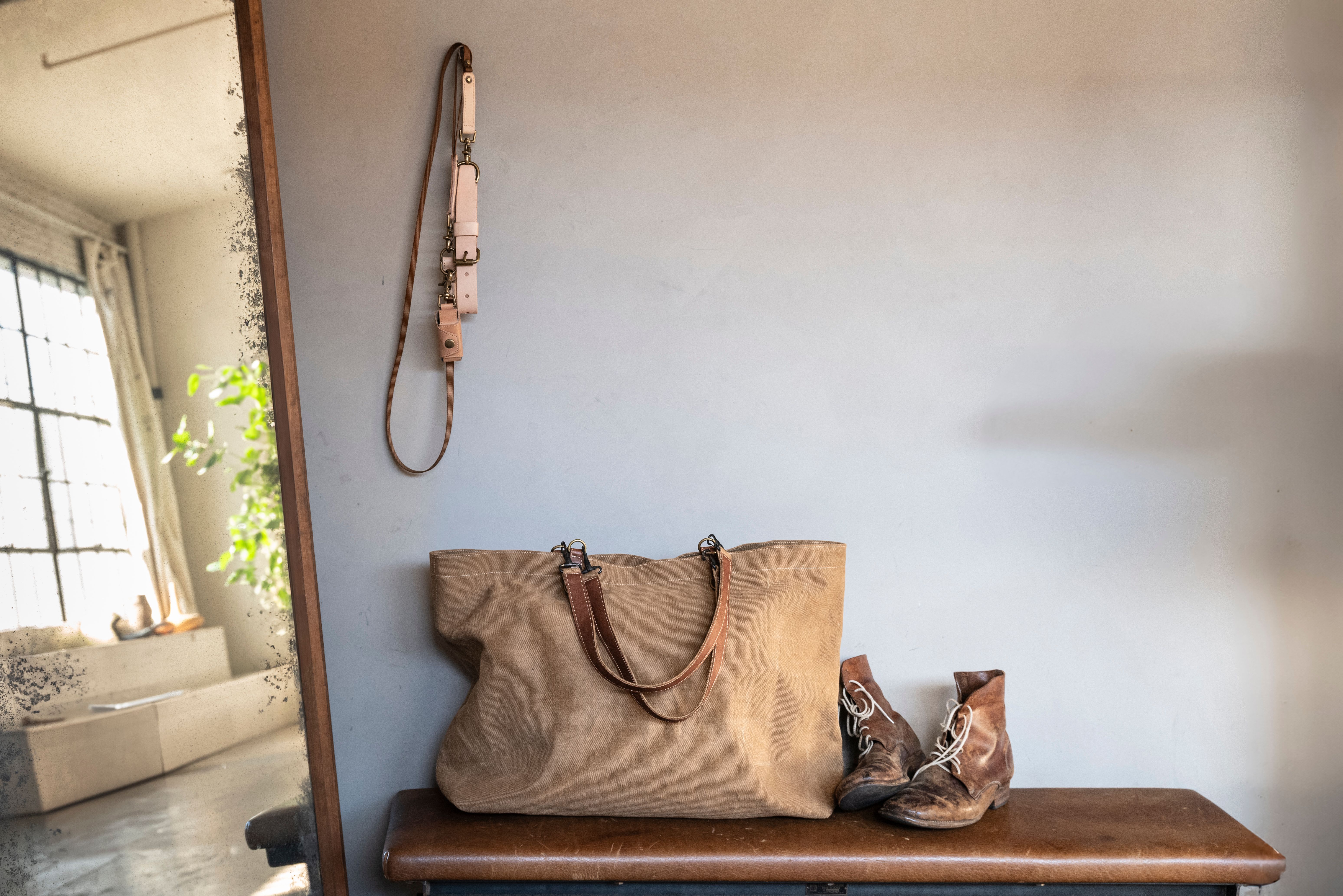 A canvas tote bag and worn boots sit on a wooden bench below a hanging leather leash and collar, beside a mirror reflecting a bright room.