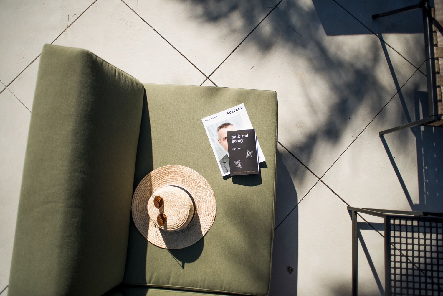 An olive green outdoor cushion with a straw hat, sunglasses, a Surface magazine, and a 'milk and honey' book on a tiled patio.
