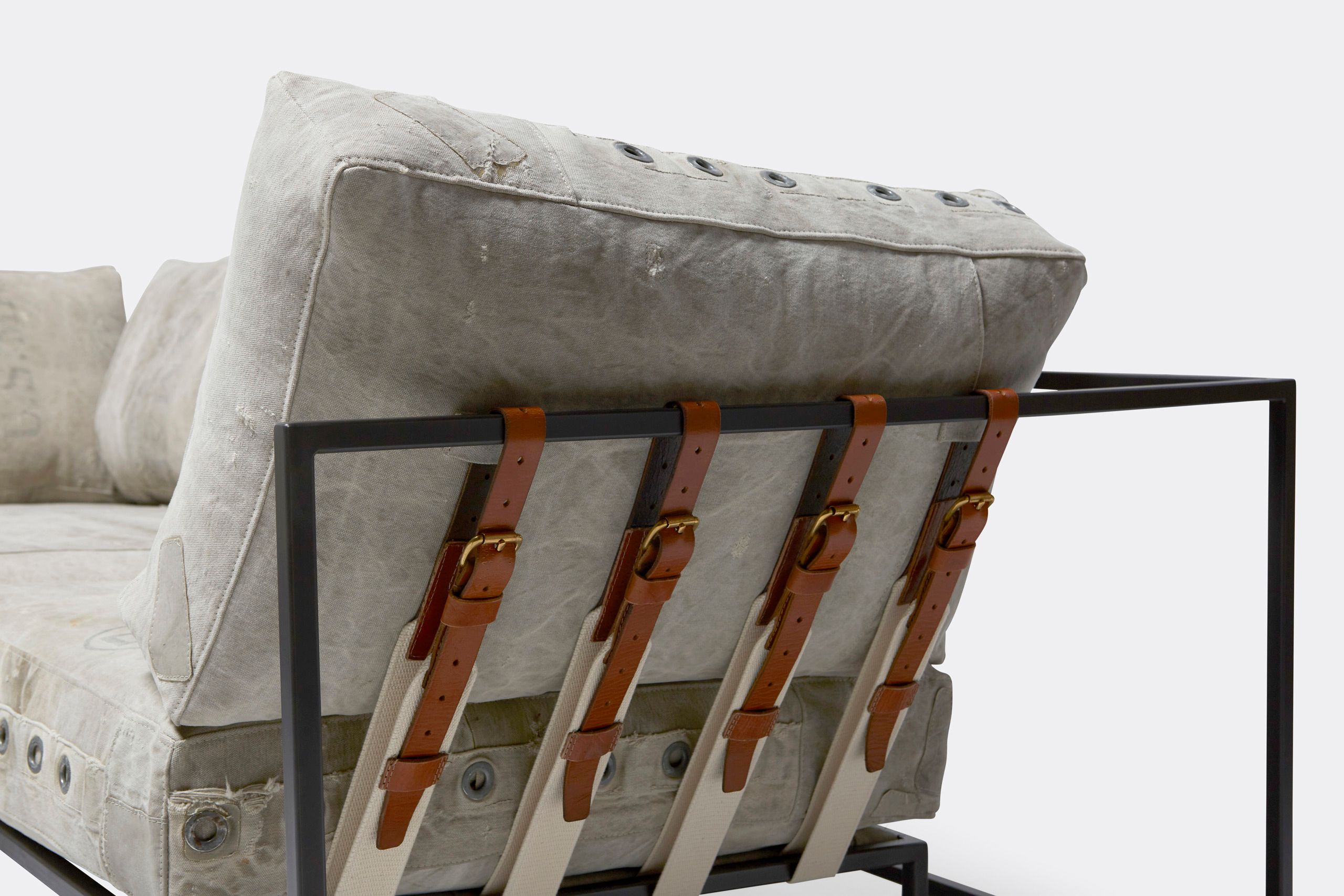 A sofa on with vintage grey canvas covers and leather and webbing straps and on a metal frame shown on a white background