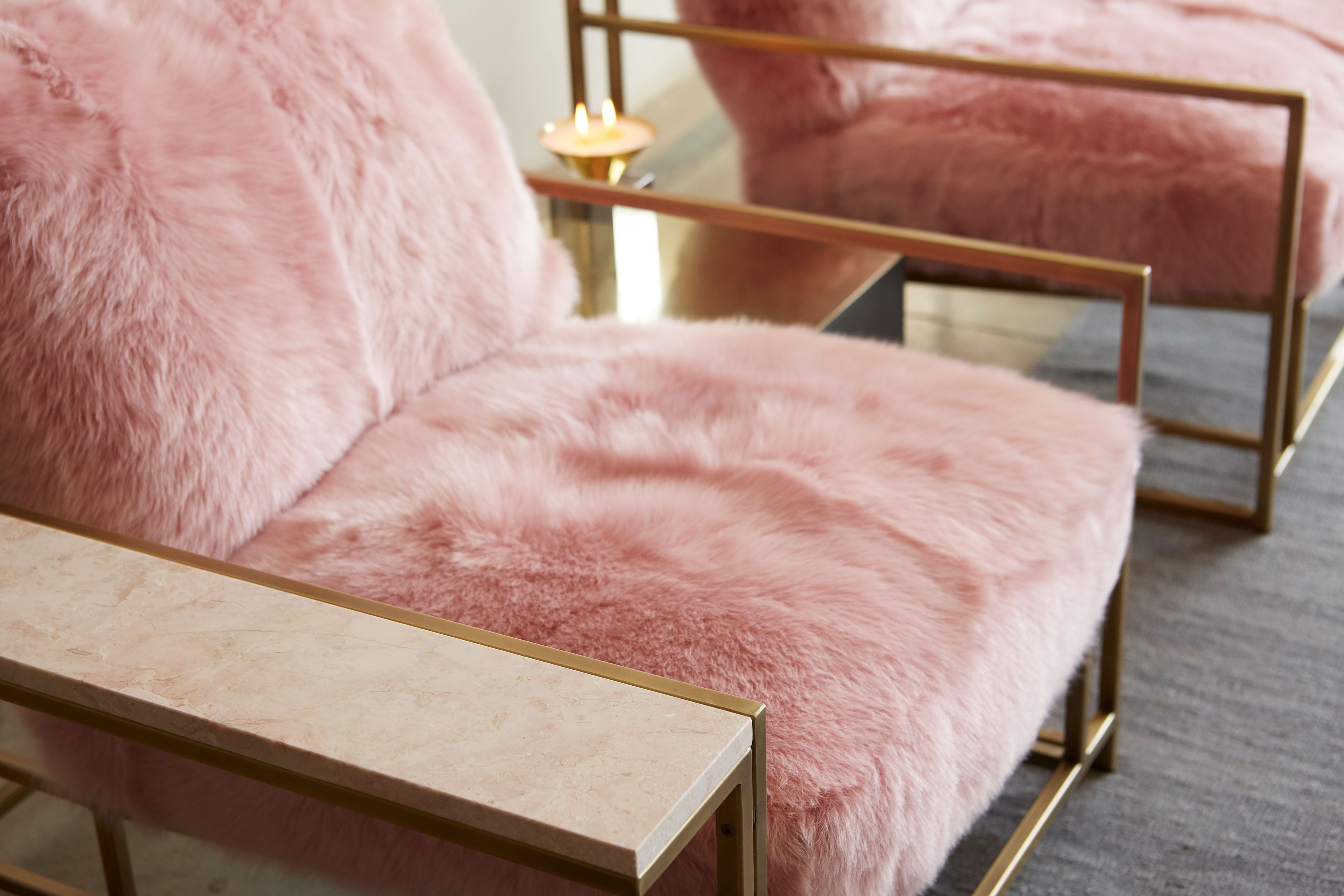 a pink shearling armchair with leather and webbing supporting belts on a brass finished steel frame and a pink marble side table attached shown on a white background .