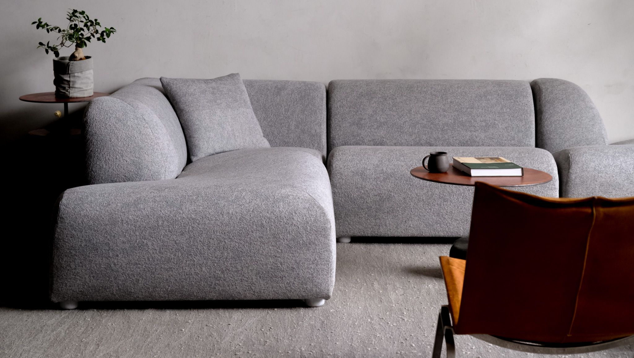 A modern gray L-shaped modular sofa in a minimalist room with side tables and a brown leather chair.